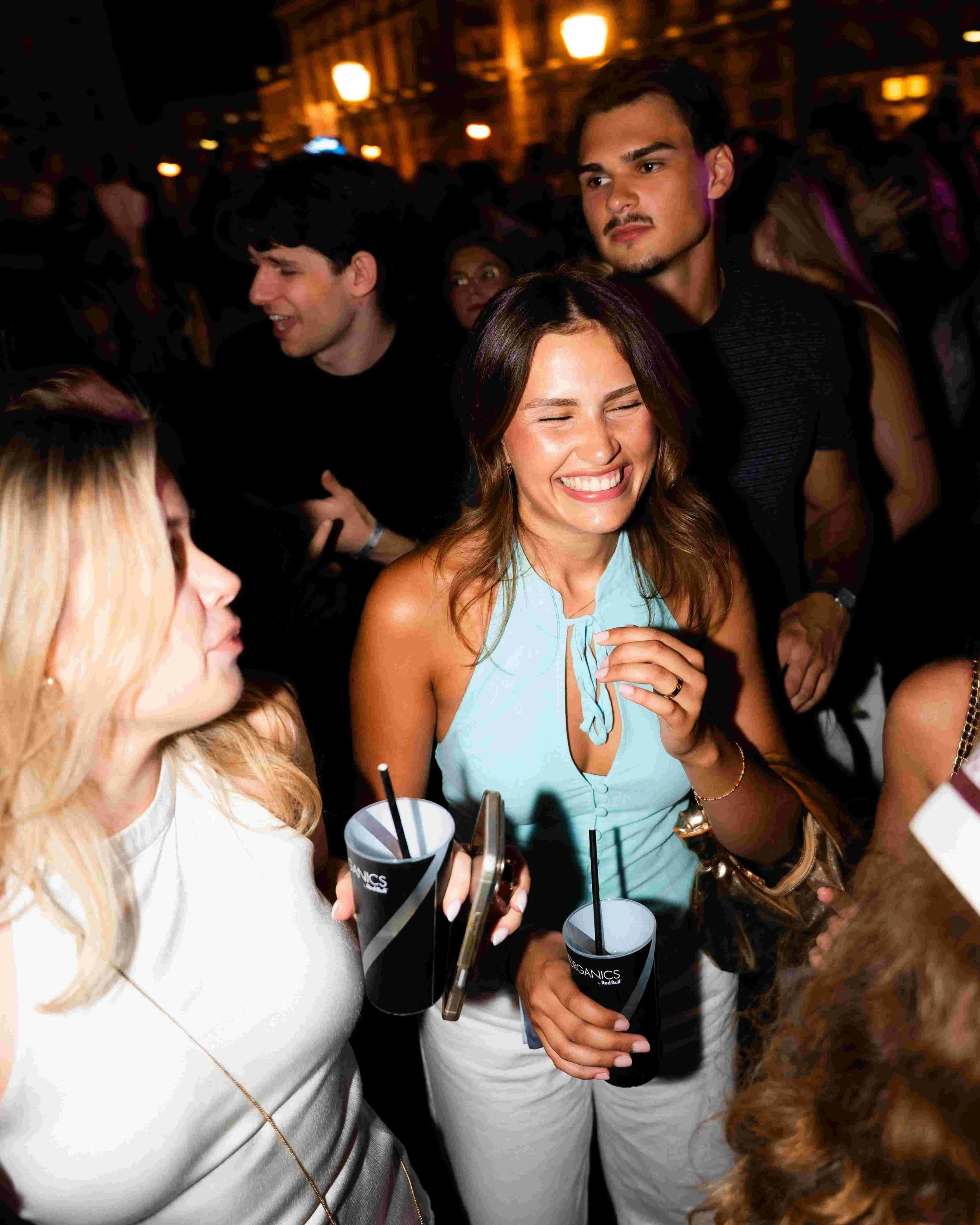 People socializing at a nighttime event in an outdoor setting, with warm lights in the background. Some are holding drinks and wearing casual summer attire.