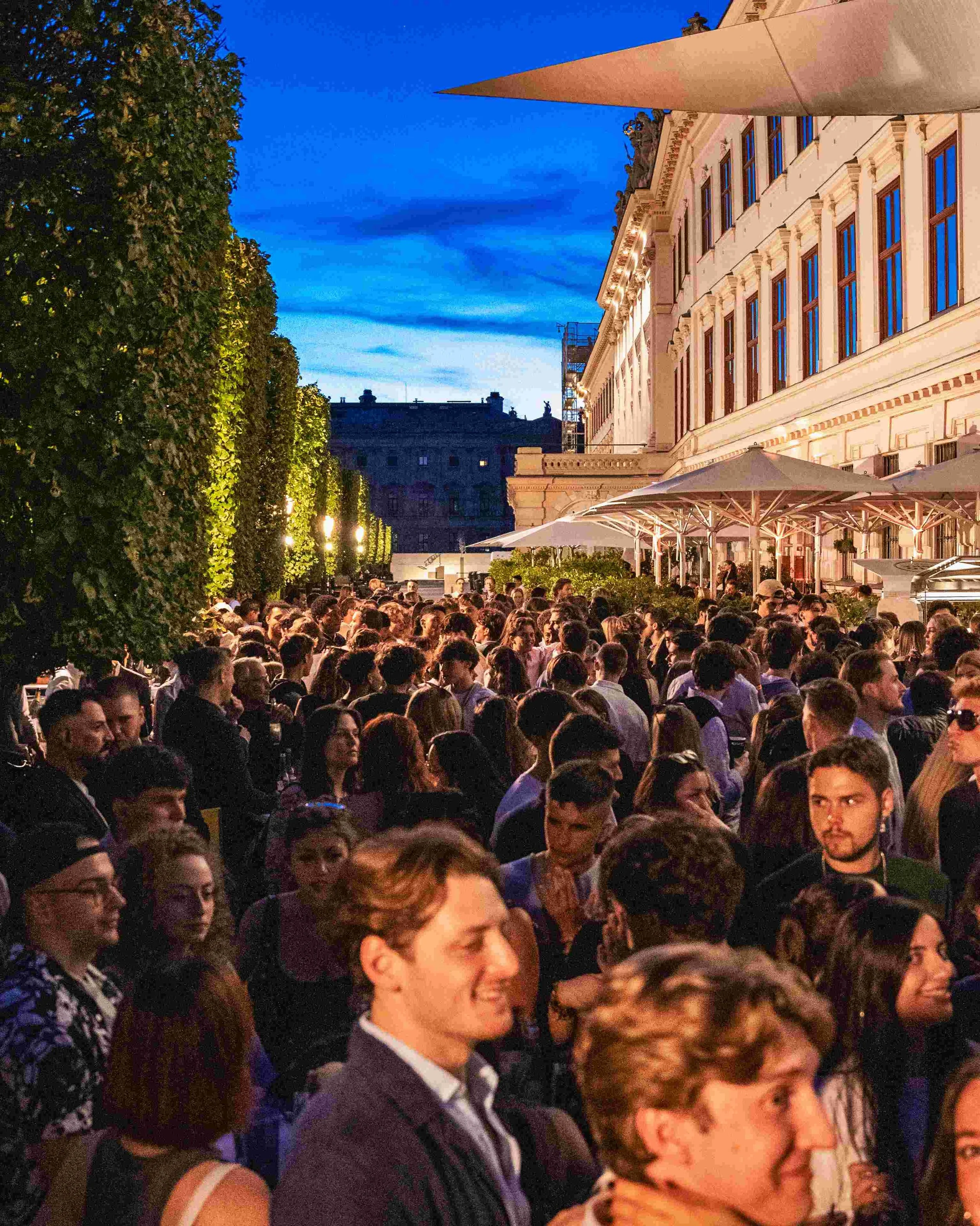 A large crowd of people gathered outdoors in front of a historic building during dusk, with some people smiling and talking, and trees illuminated by lights on the left.