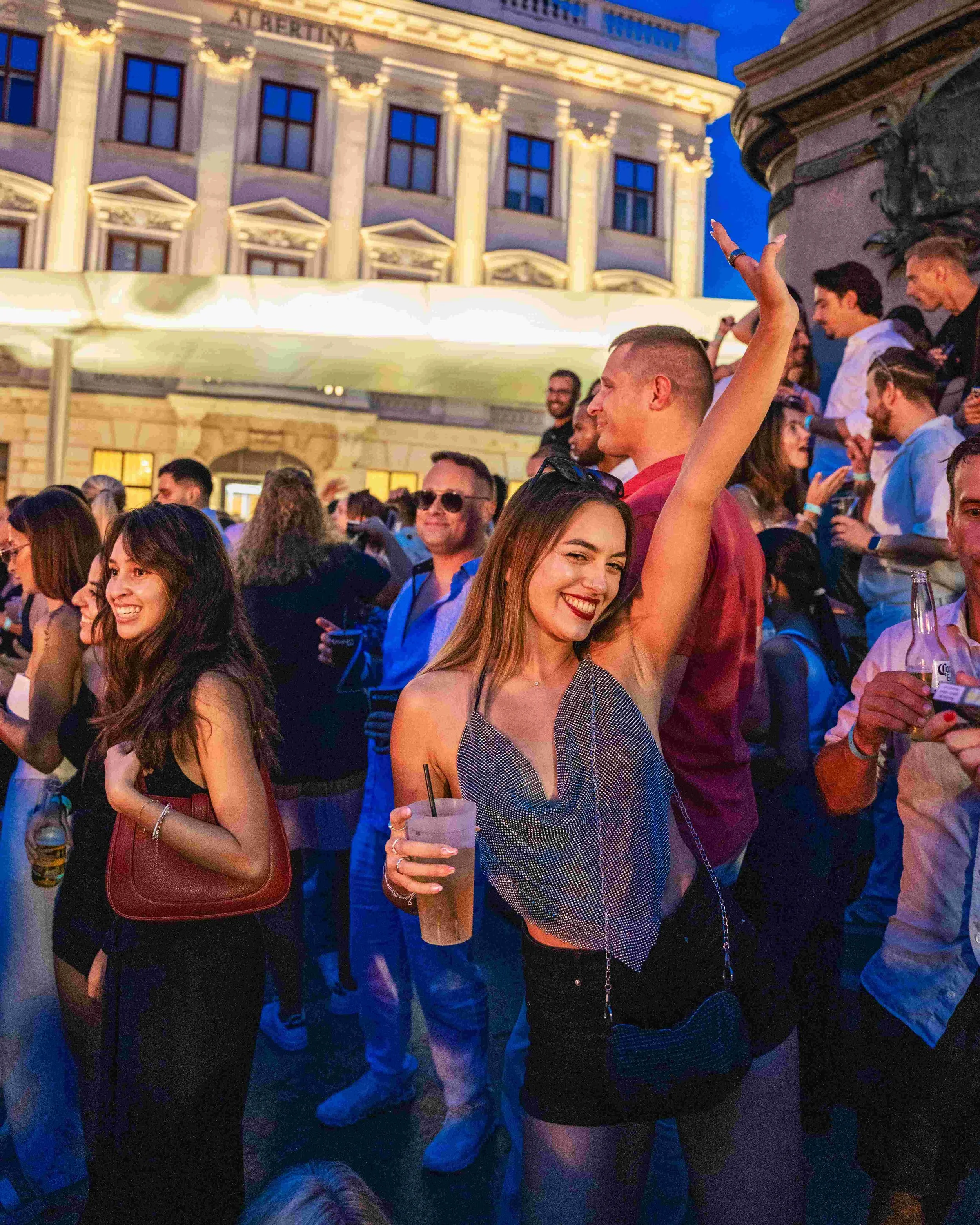 Crowd of people socializing outdoors at night, with a historic building illuminated in the background.