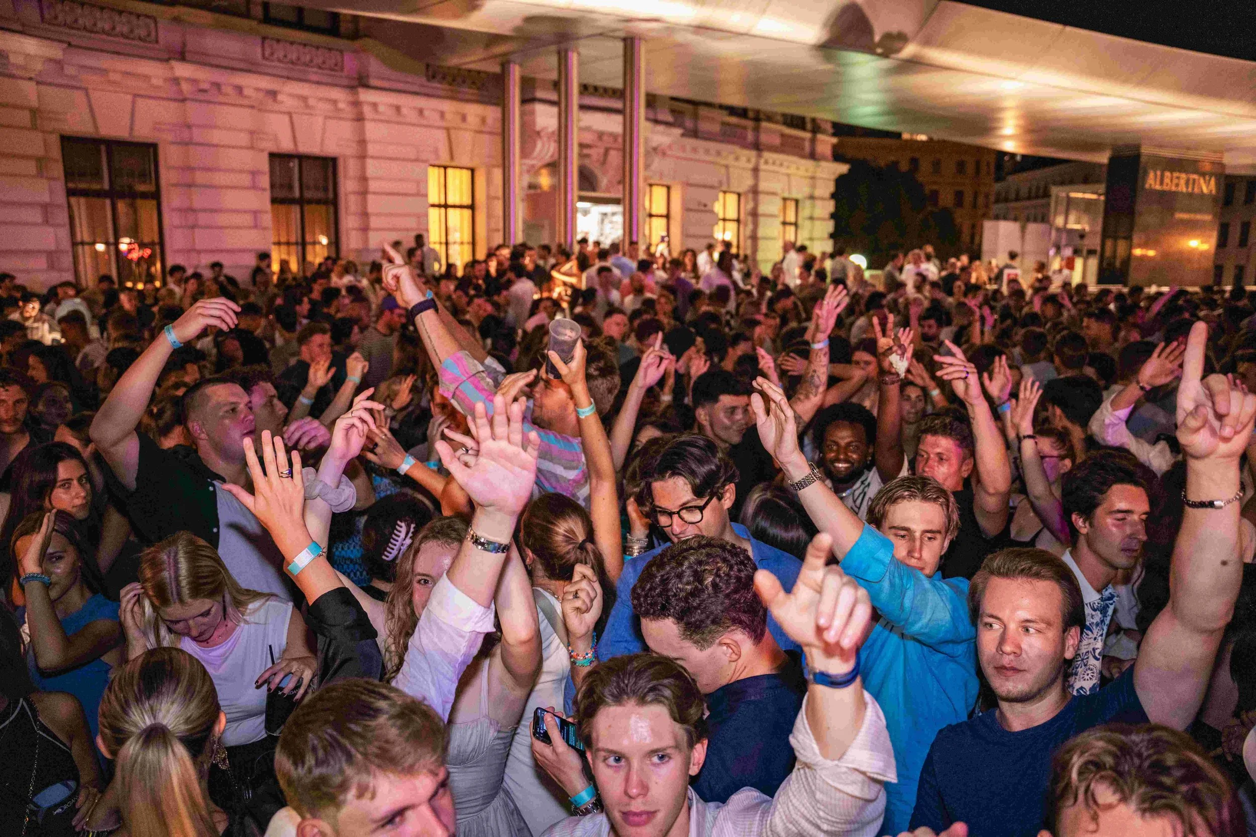 A large crowd of people dancing and enjoying a nighttime event on a balcony with a pink building in the background.
