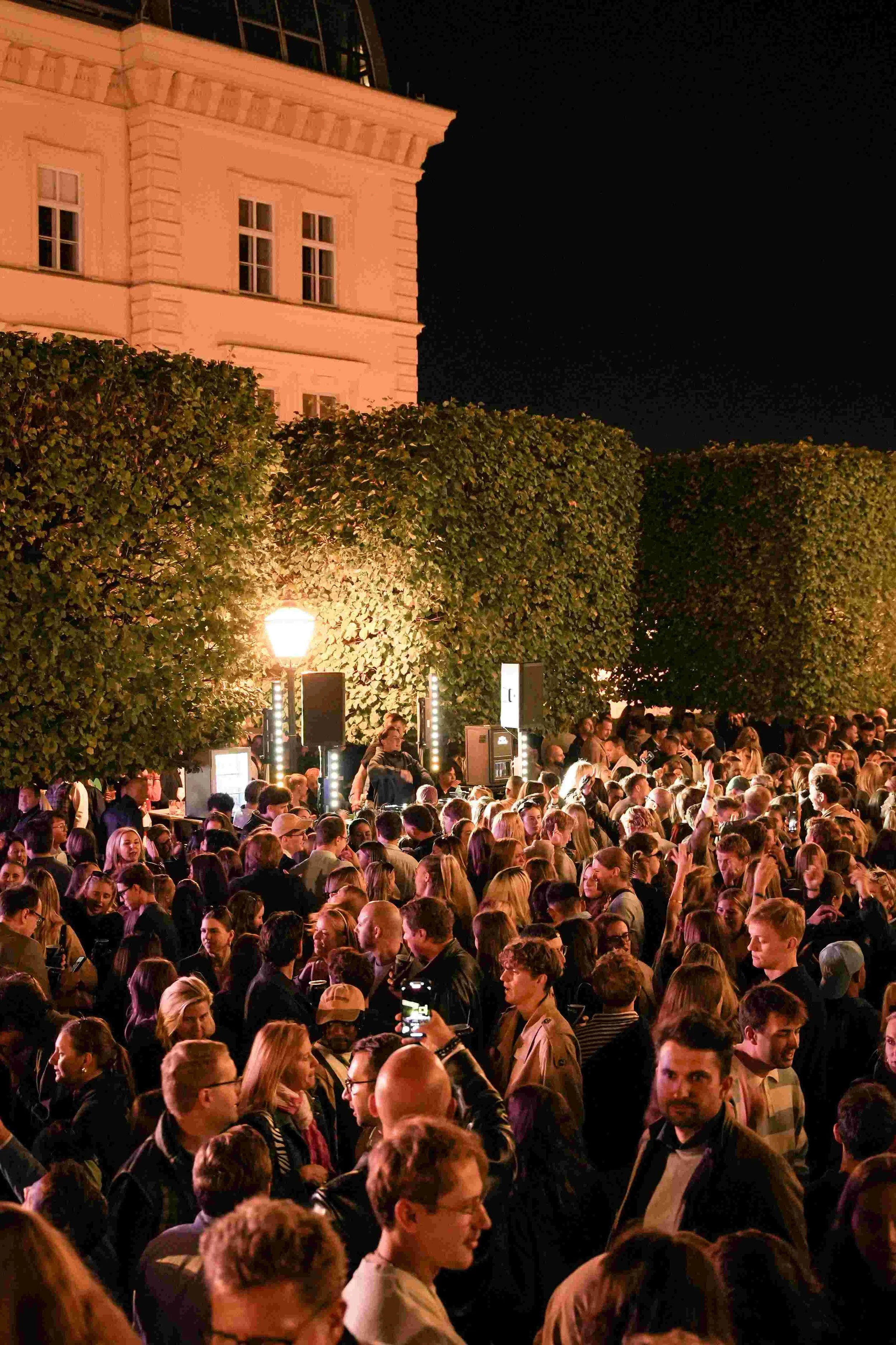 Crowd of people gathered outdoors at night, illuminated by streetlights, with a building and dark sky in the background.