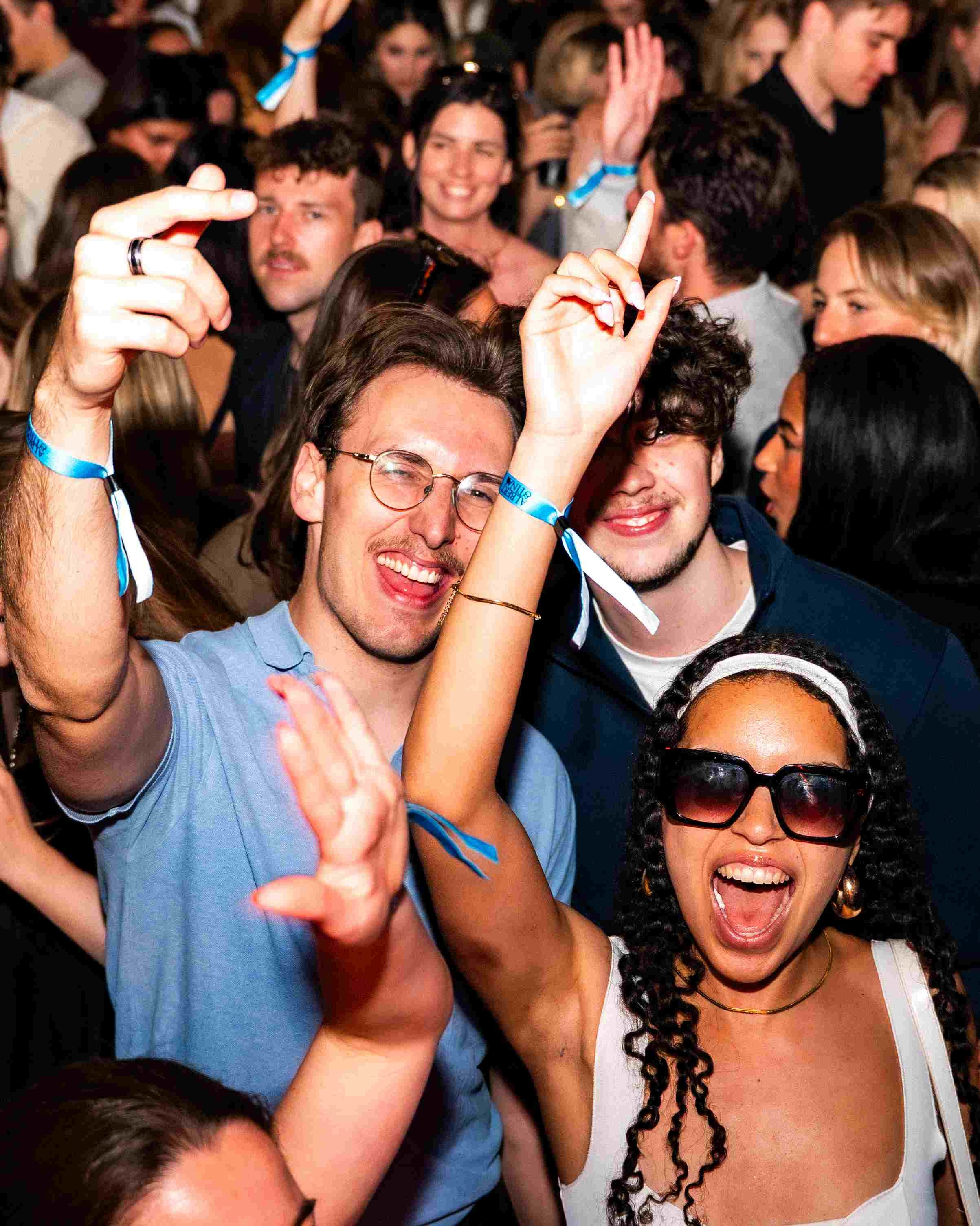 A large group of young people at a lively party or celebration, smiling, dancing, and making gestures with their hands, including peace signs and raised fingers. They are wearing casual clothing and accessories, with some wearing sunglasses and wrist