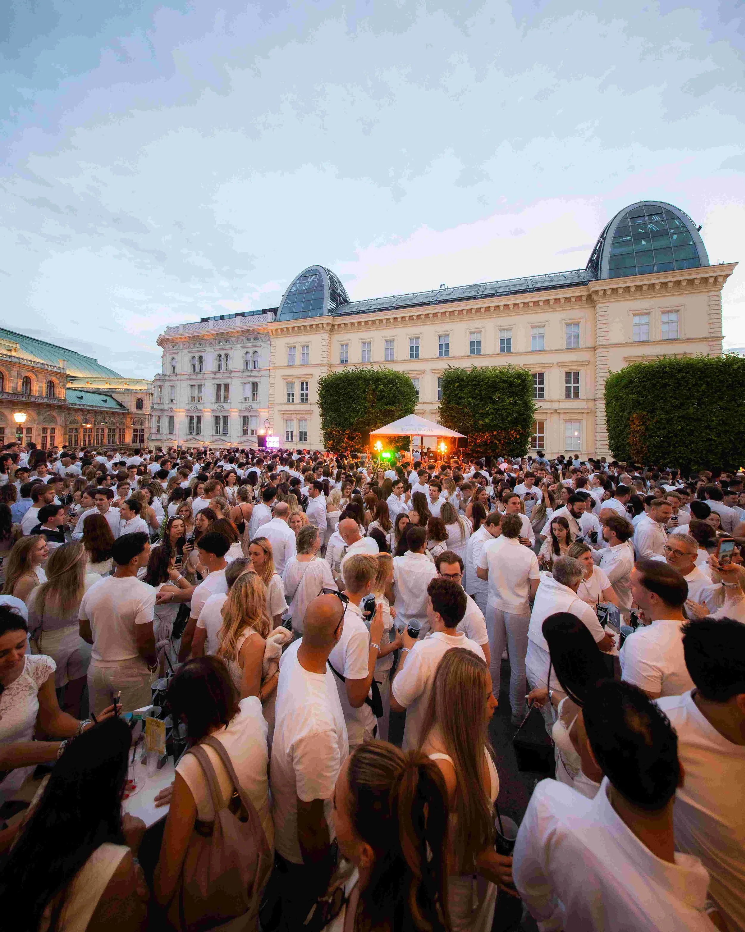 A large crowd of people gathered outdoors in front of a historic building with modern arched glass rooftops, during an evening event with stage lighting.