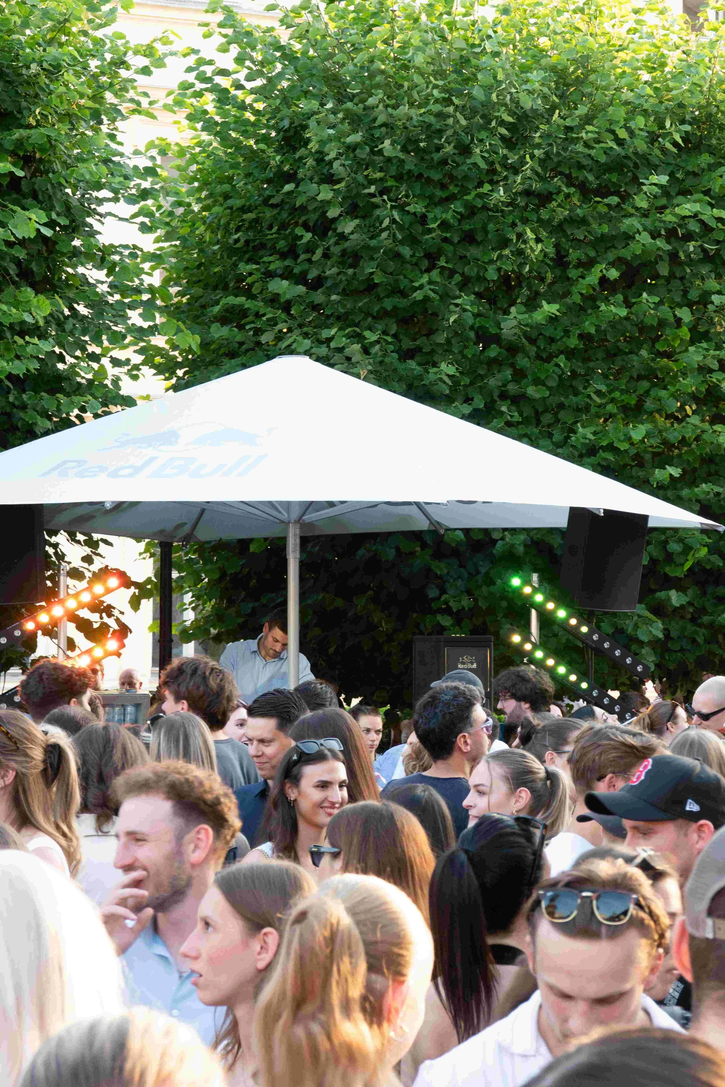 A crowded outdoor event with a diverse group of people standing close together. There is a stage with musical or DJ equipment, lights, speakers, and a large white umbrella with the Red Bull logo. The background features green trees and some structure