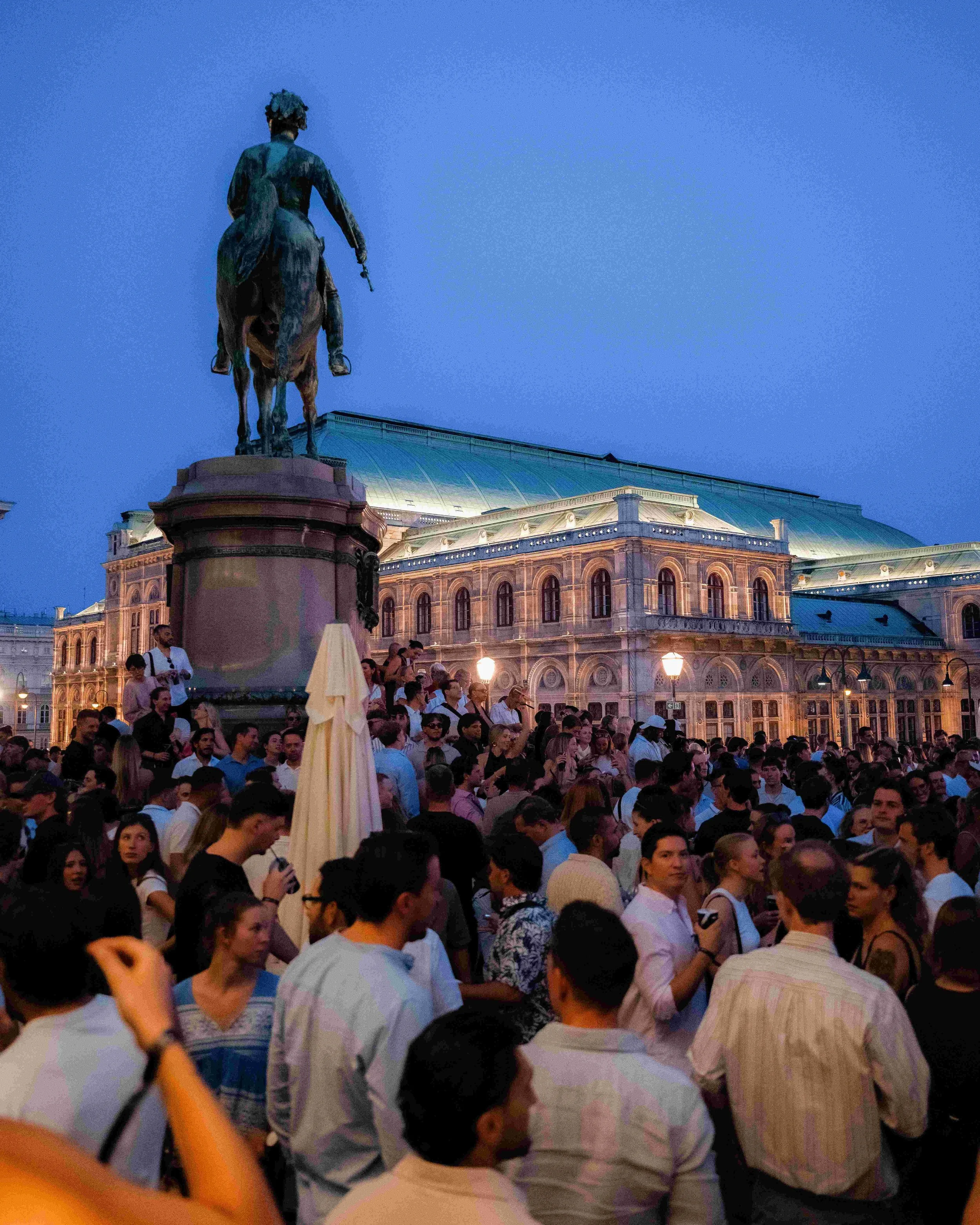Crowd gathered at dusk in front of a historic building with a large statue of a mounted figure on a pedestal.