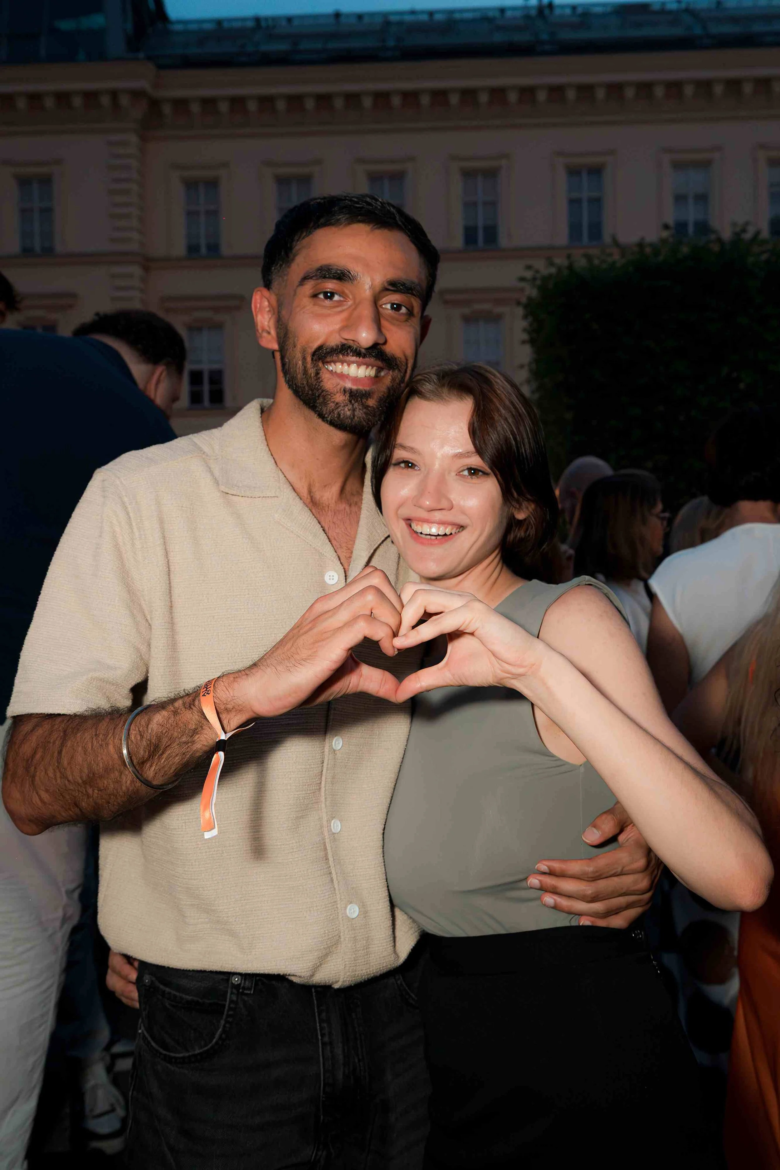 A smiling man and woman holding hands together in a heart shape, at an outdoor event during evening, with a crowd and building in the background.