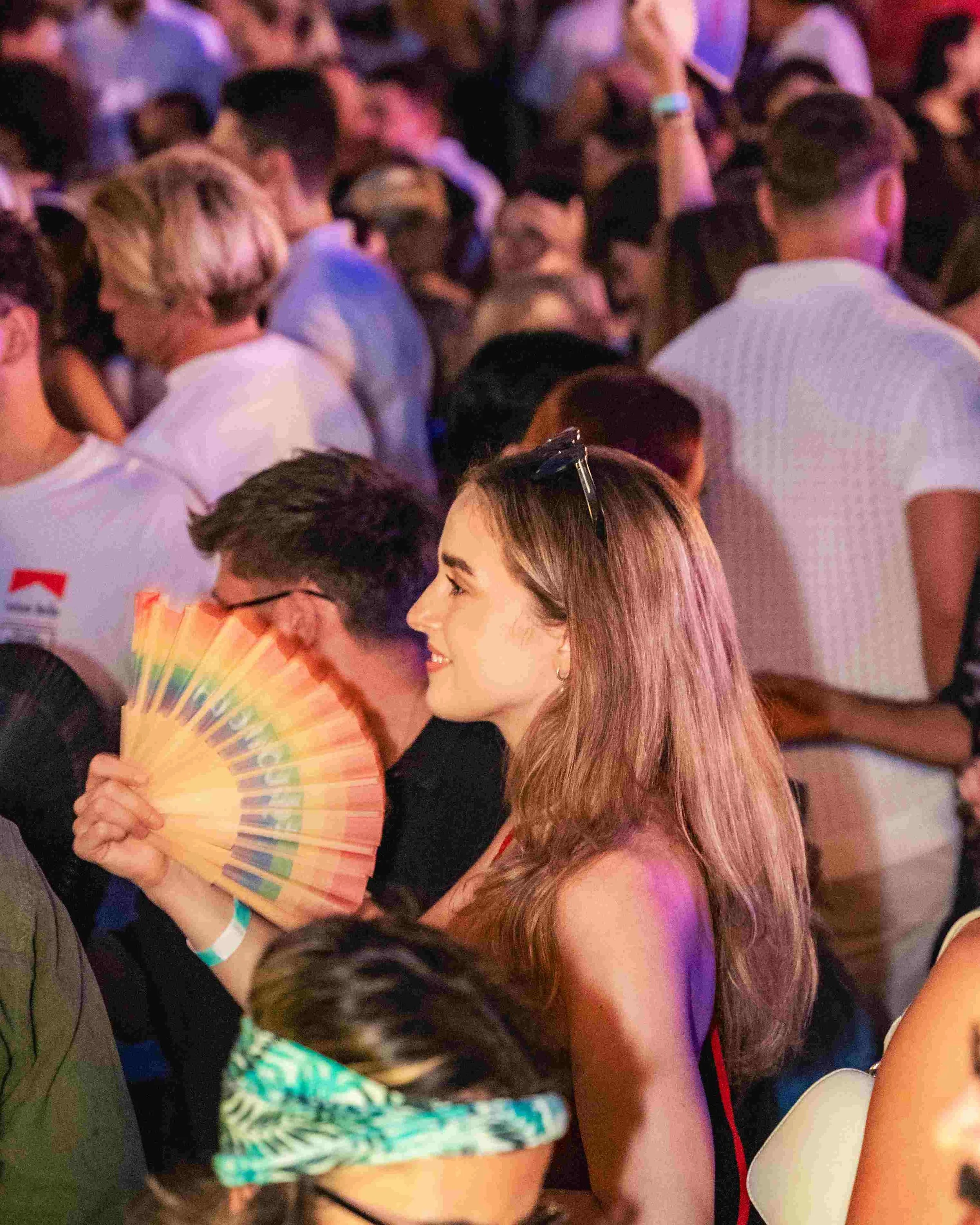 A crowded group of people at a party or social event, some holding drinks and a fan, with colorful lighting.
