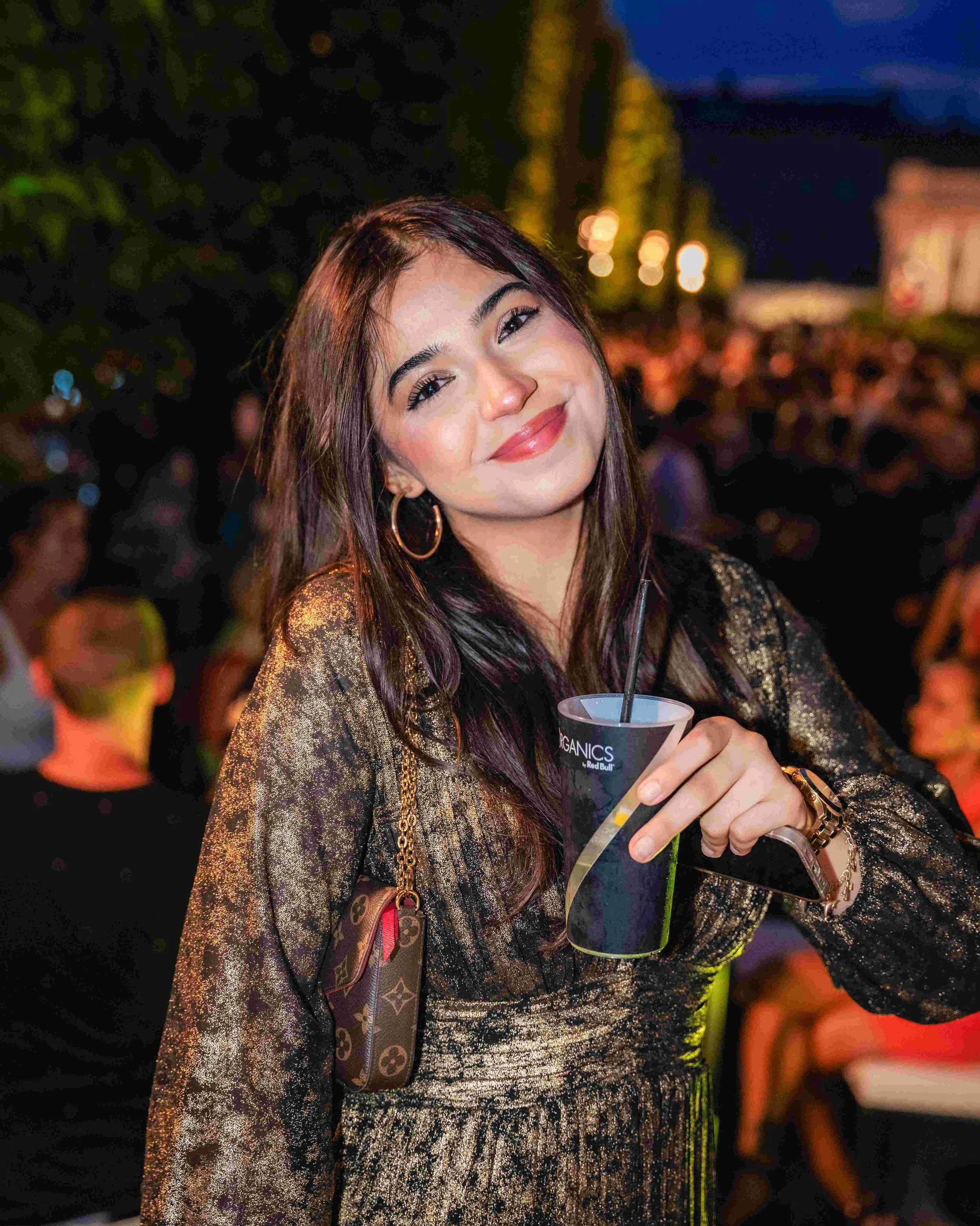 A woman with long brown hair, wearing gold hoop earrings and a shiny gold and black patterned top, smiling while holding a drink with a black straw at a festive event during the evening or night.