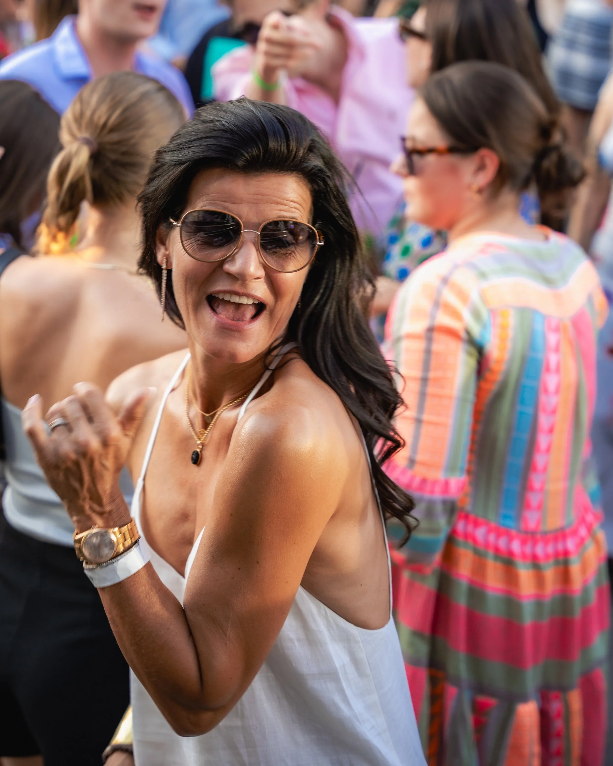 A woman with dark hair, wearing sunglasses, a gold watch, and jewelry, smiling and reaching her hand out at an outdoor event with multiple people in colorful clothing around her.