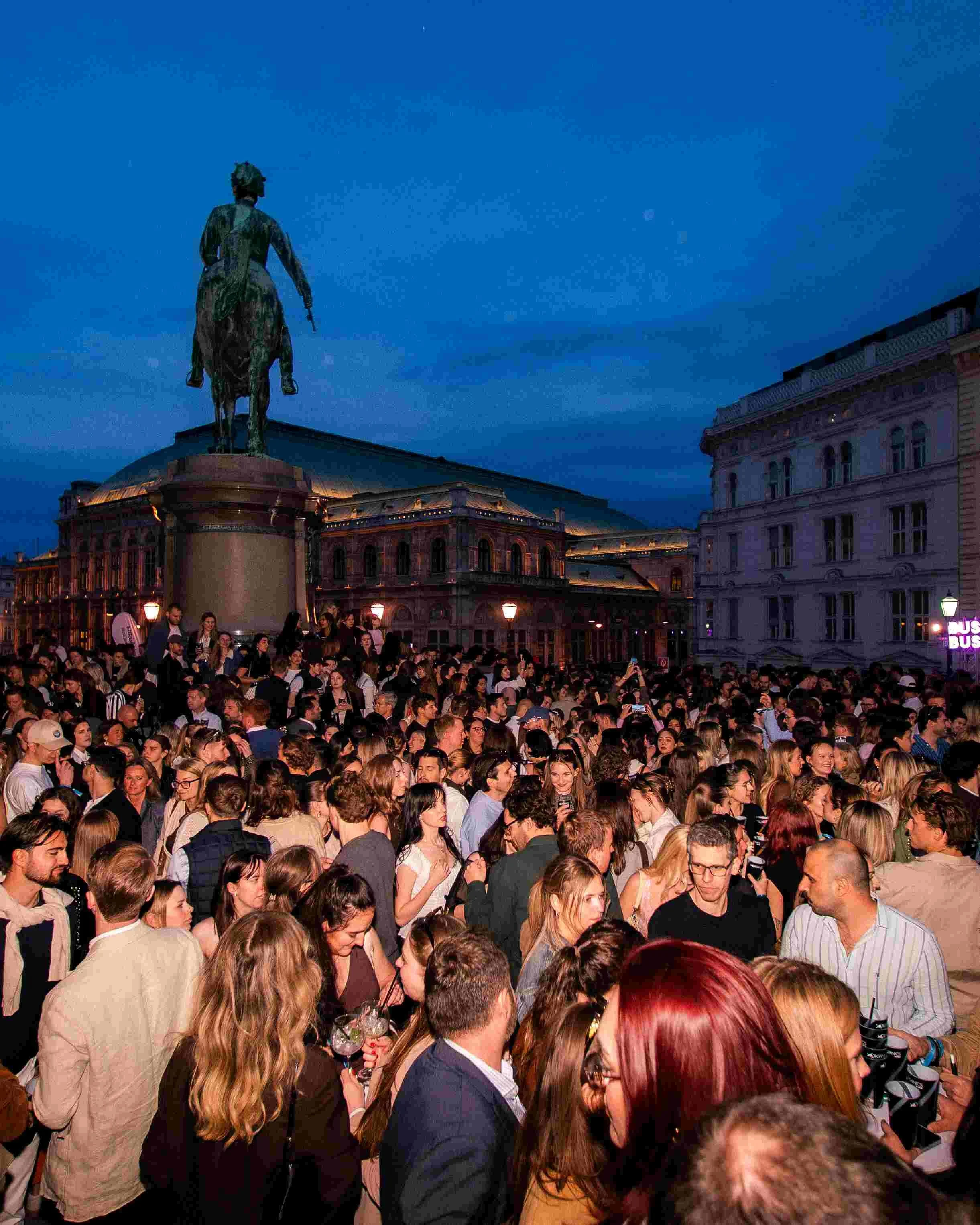 A large crowd of people gathered outside, with a statue of a person on horseback on a pedestal in the background during the evening. Buildings and streetlights are also visible.