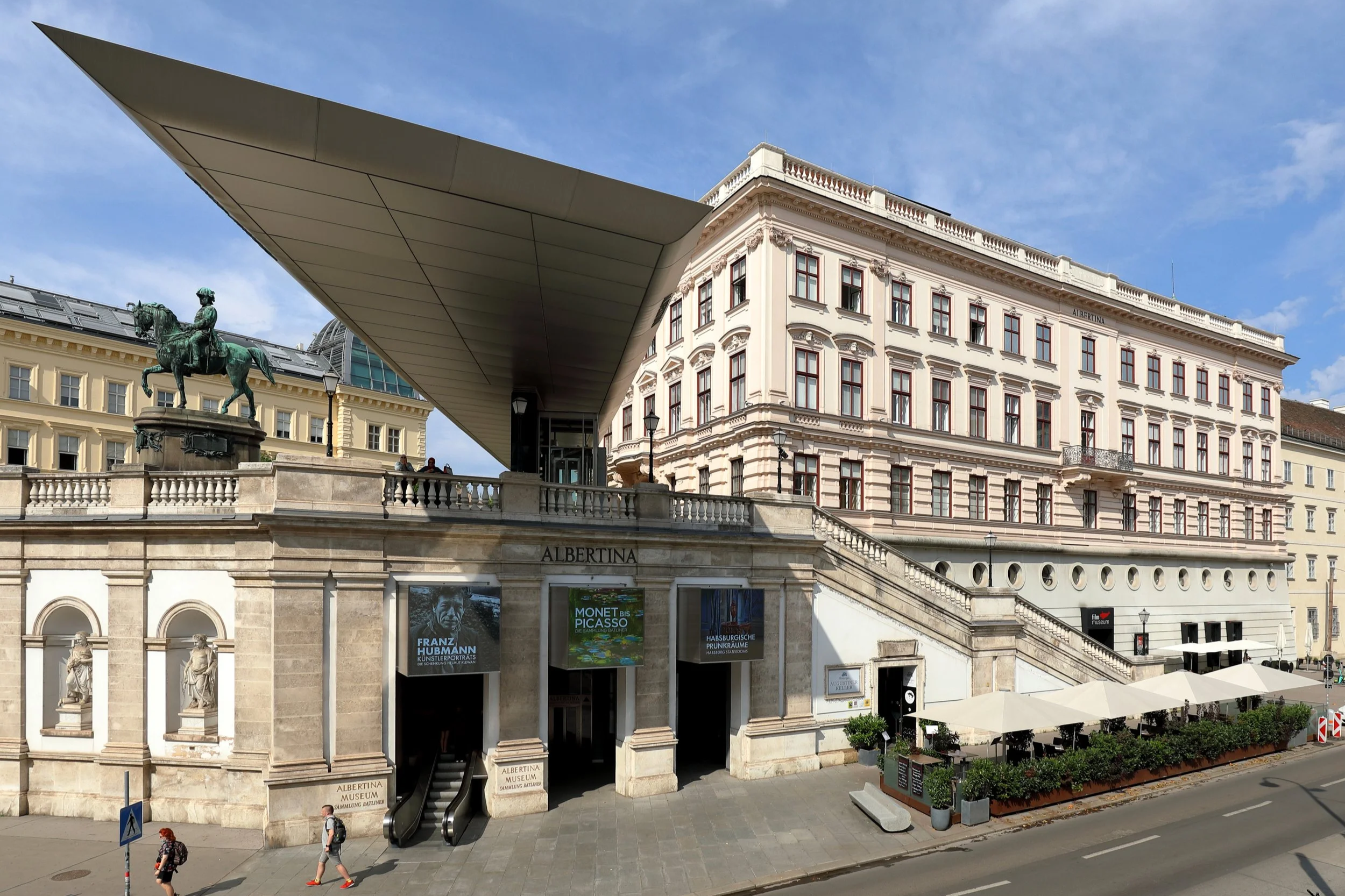 The Albertina Museum building in Vienna, Austria, with a modern overhanging roof, classical architecture, a statue of a man on a horse, and people walking by.