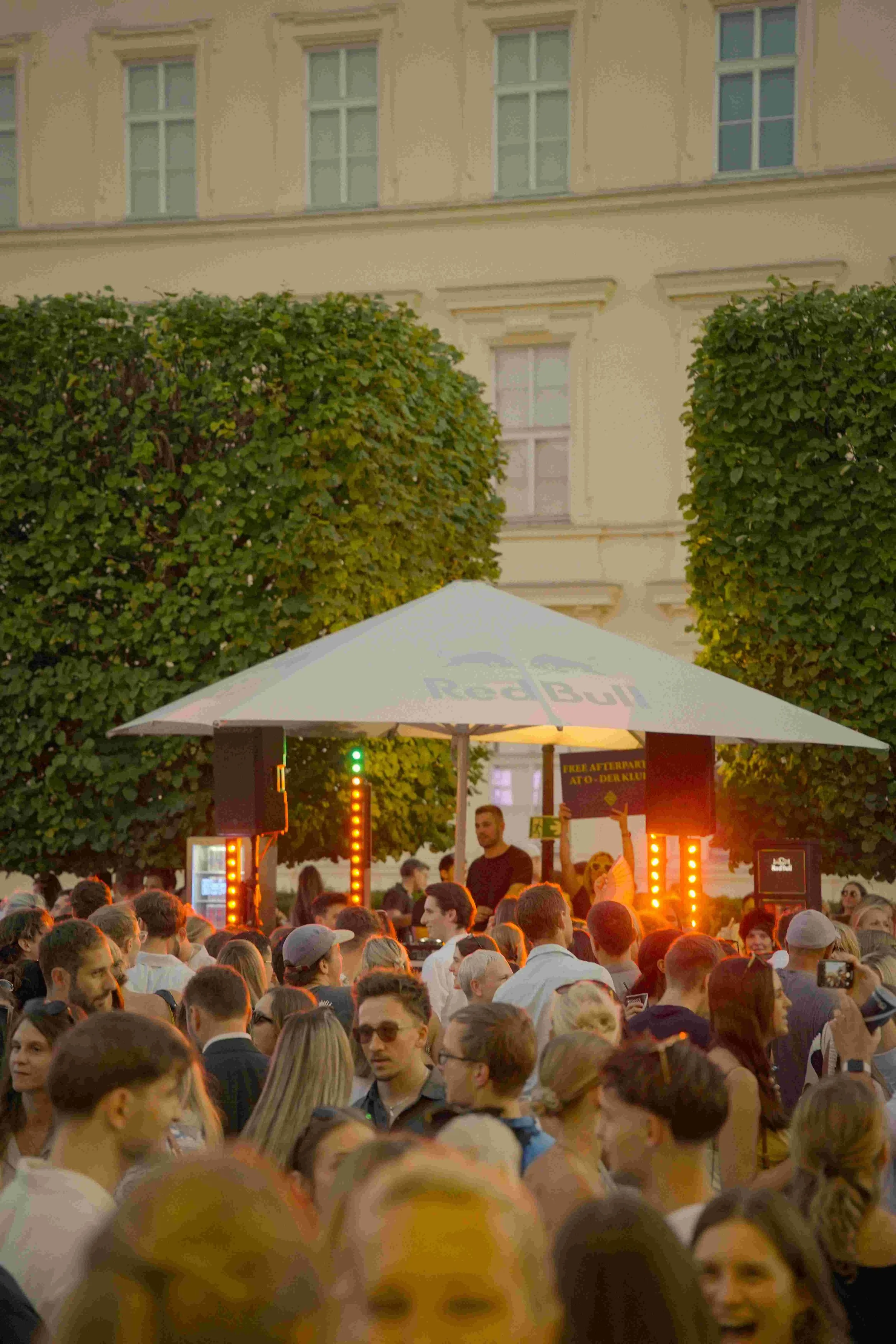 Crowd of people gathered outdoors at a concert or event during sunset, with trees and a building in the background.