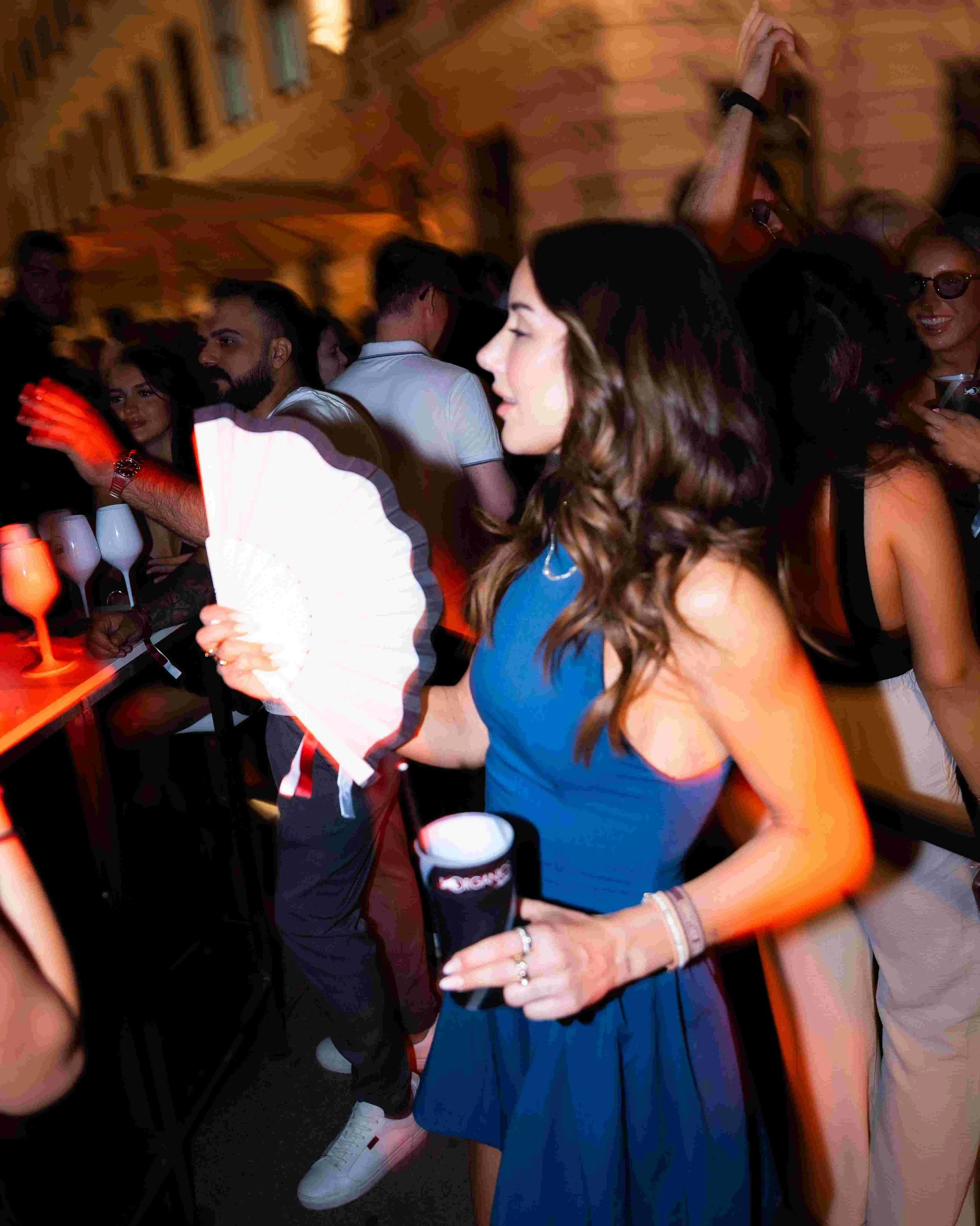 People dancing at a party or celebration, some holding drinks, with a woman in a blue dress as a main focus.
