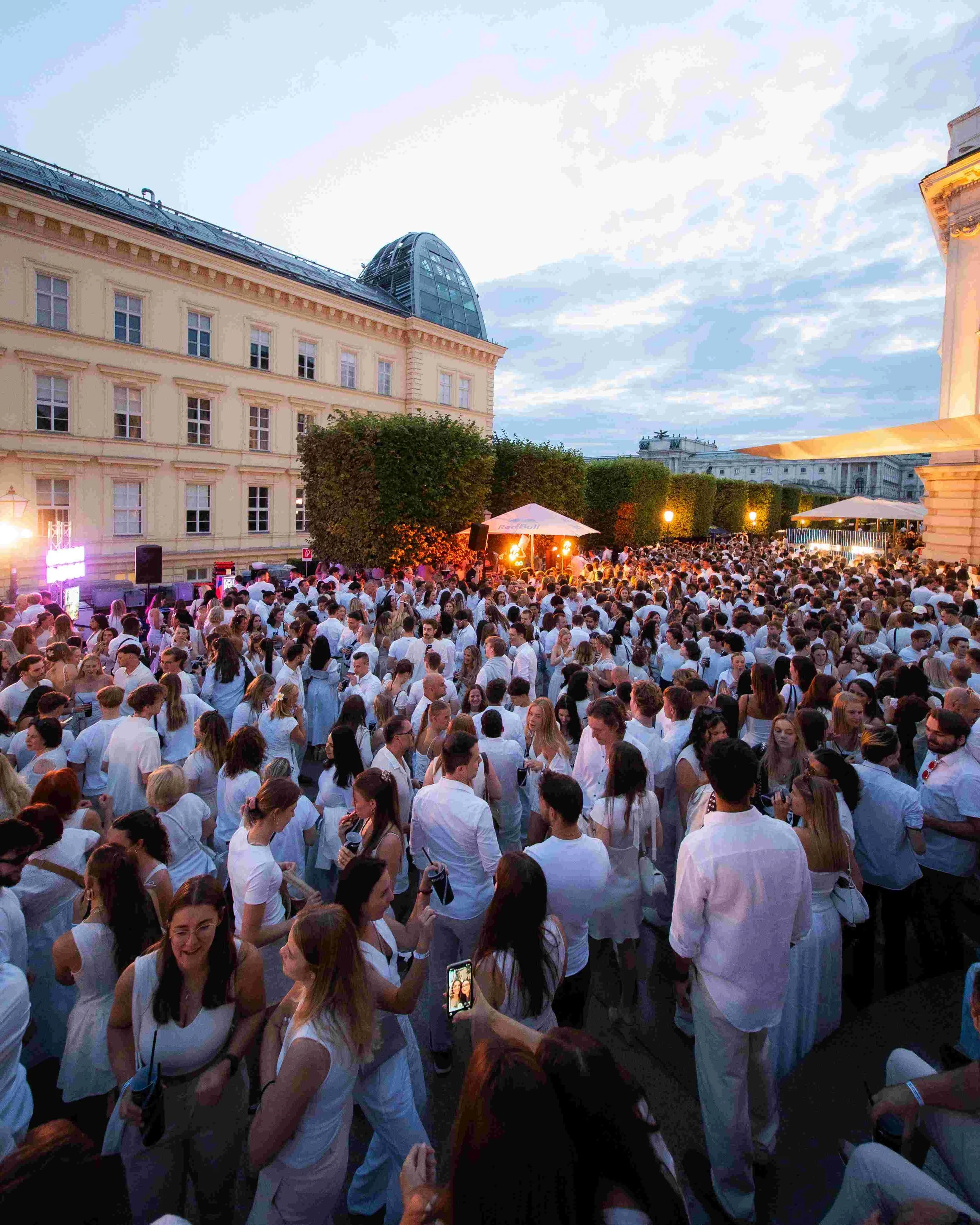 Crowd of people dressed in white at an outdoor event during sunset, with historic buildings and trees in the background.