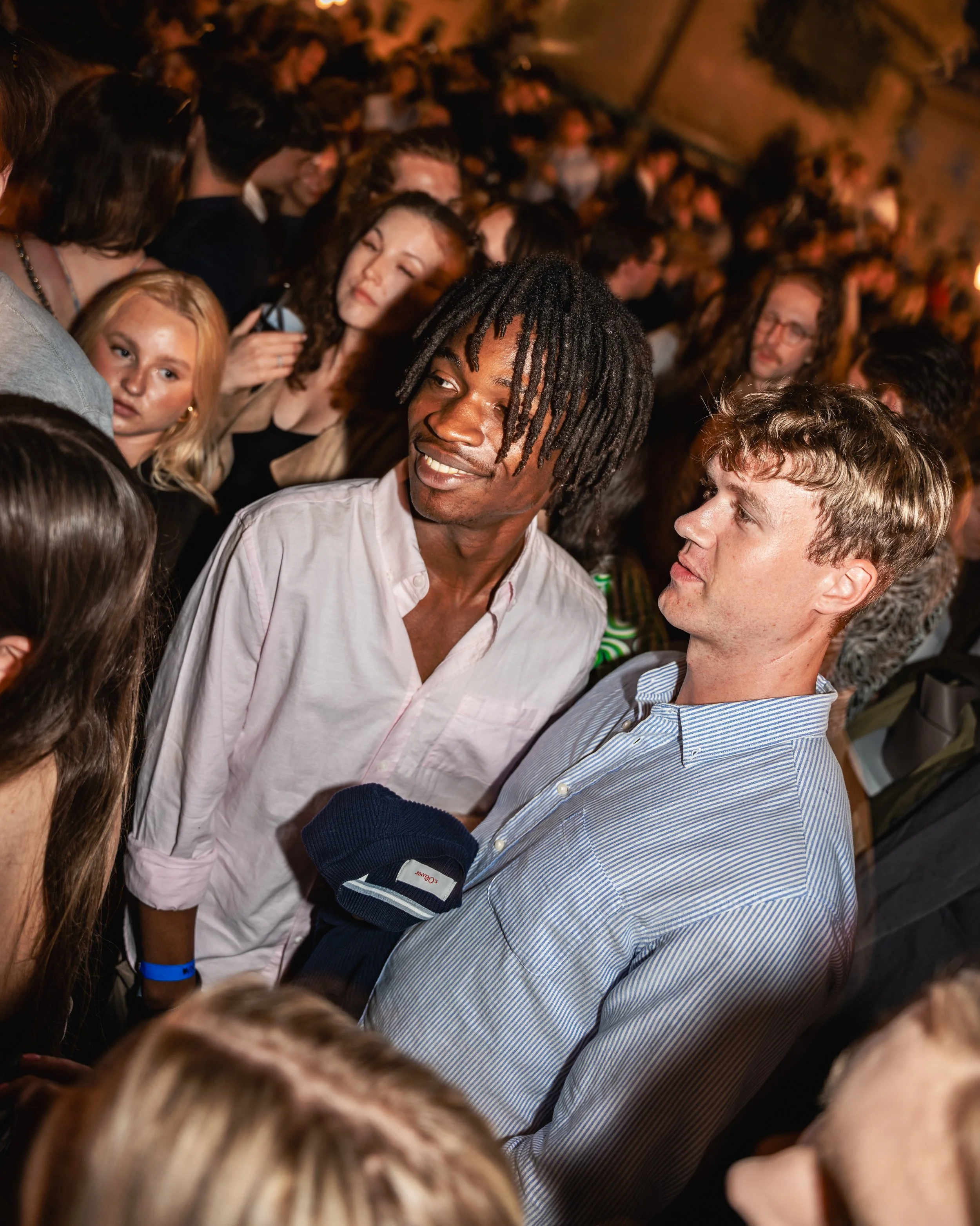A crowd of people at a social gathering, with two men in the foreground talking to each other. The background is filled with more people, some smiling and engaging in conversations, under warm lighting.