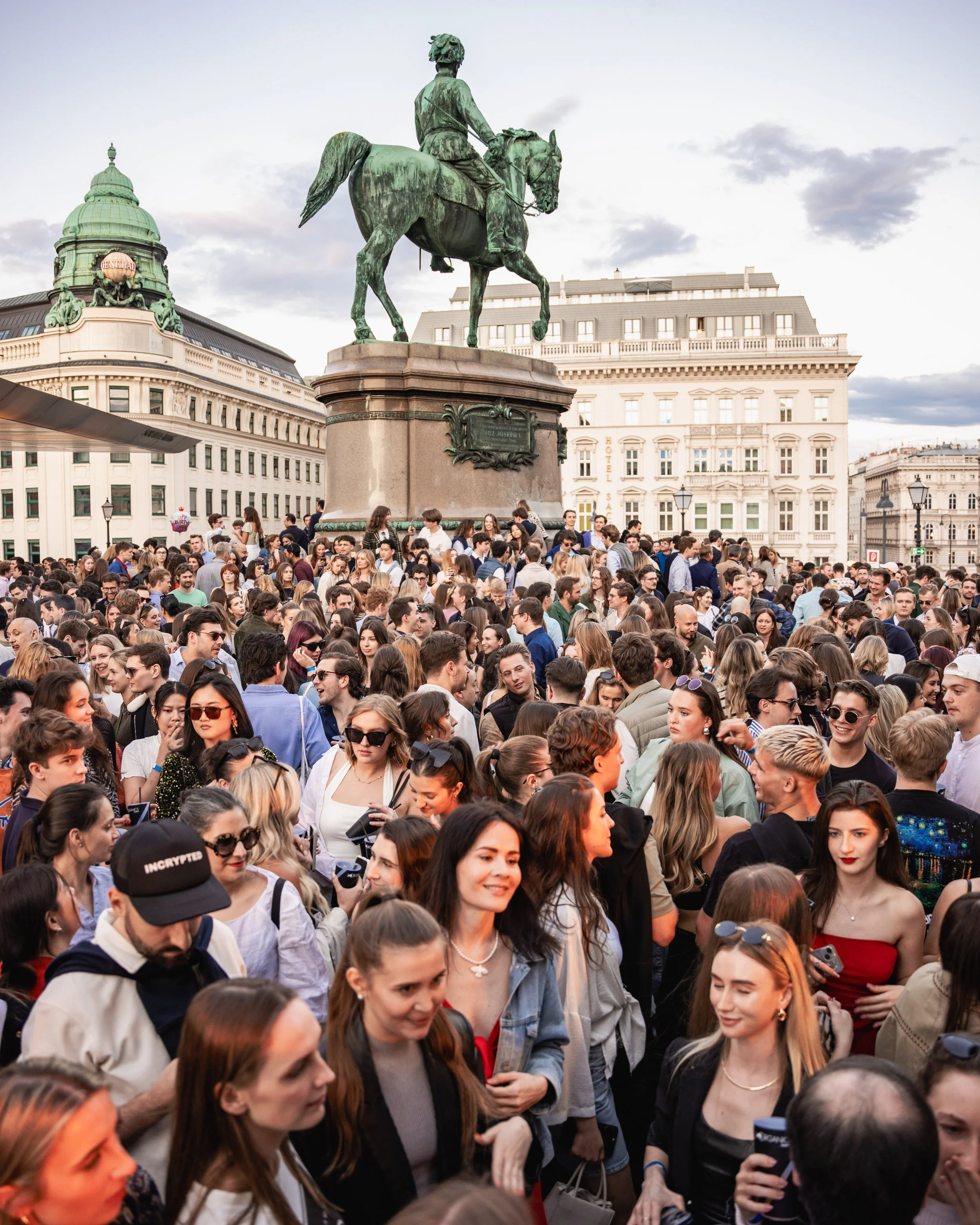 A crowded public square with a bronze equestrian statue of a man on a horse, and historic buildings in the background, including one with a prominent green dome.