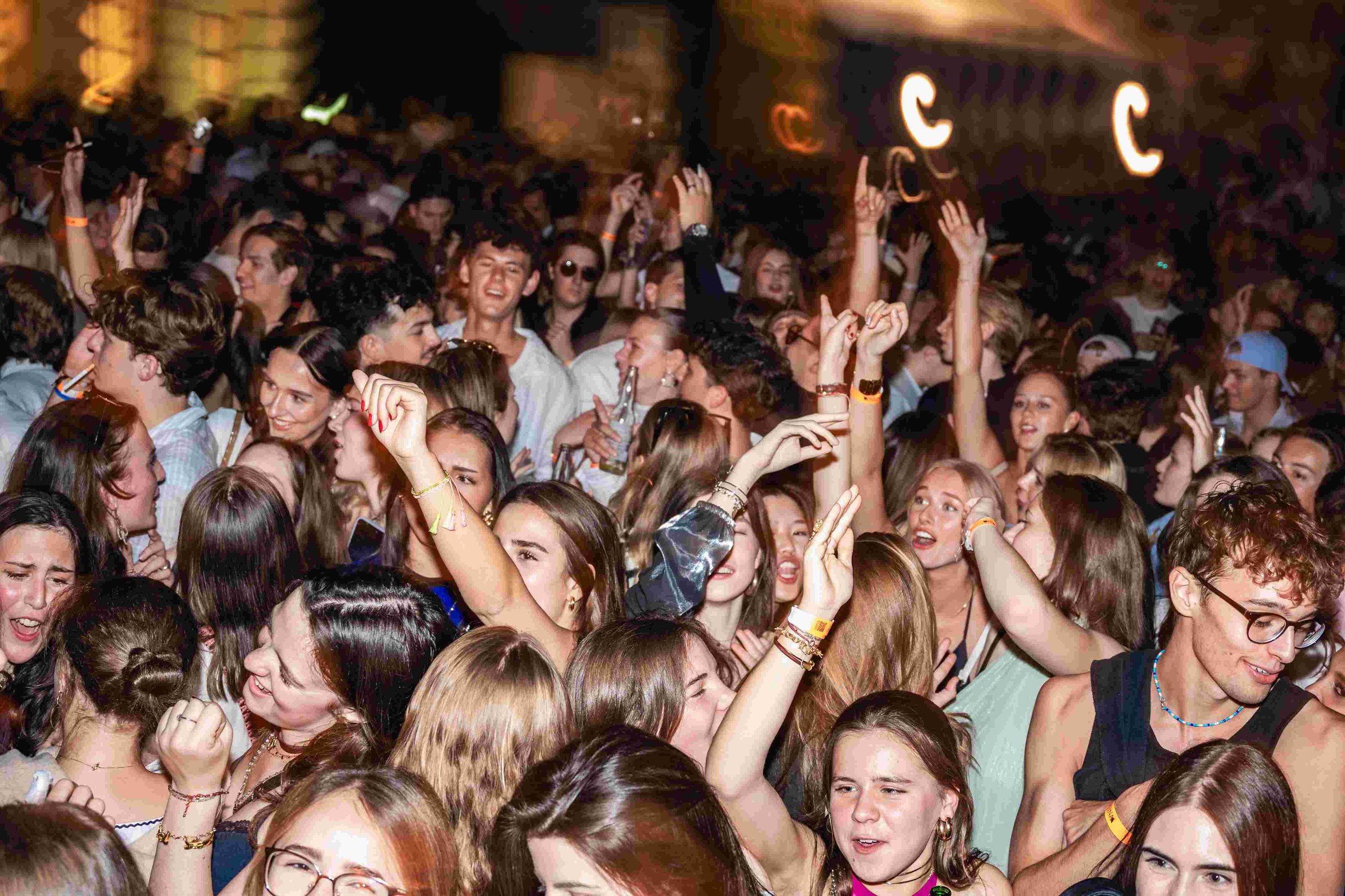 A large crowd of young people at a concert or party, dancing and raising their hands in the air.