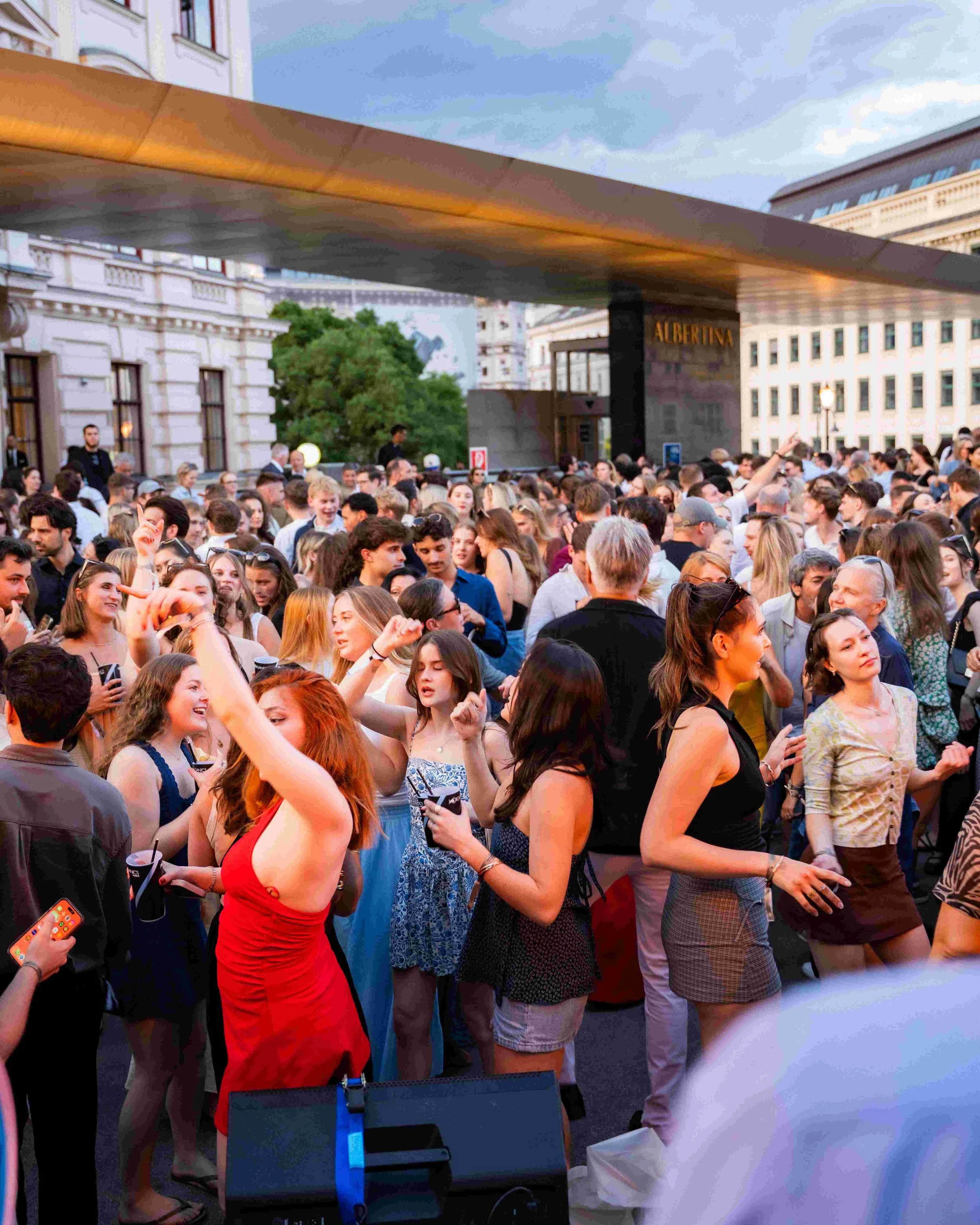 Crowd of people gathered outdoors during the evening, with a building and trees in the background.