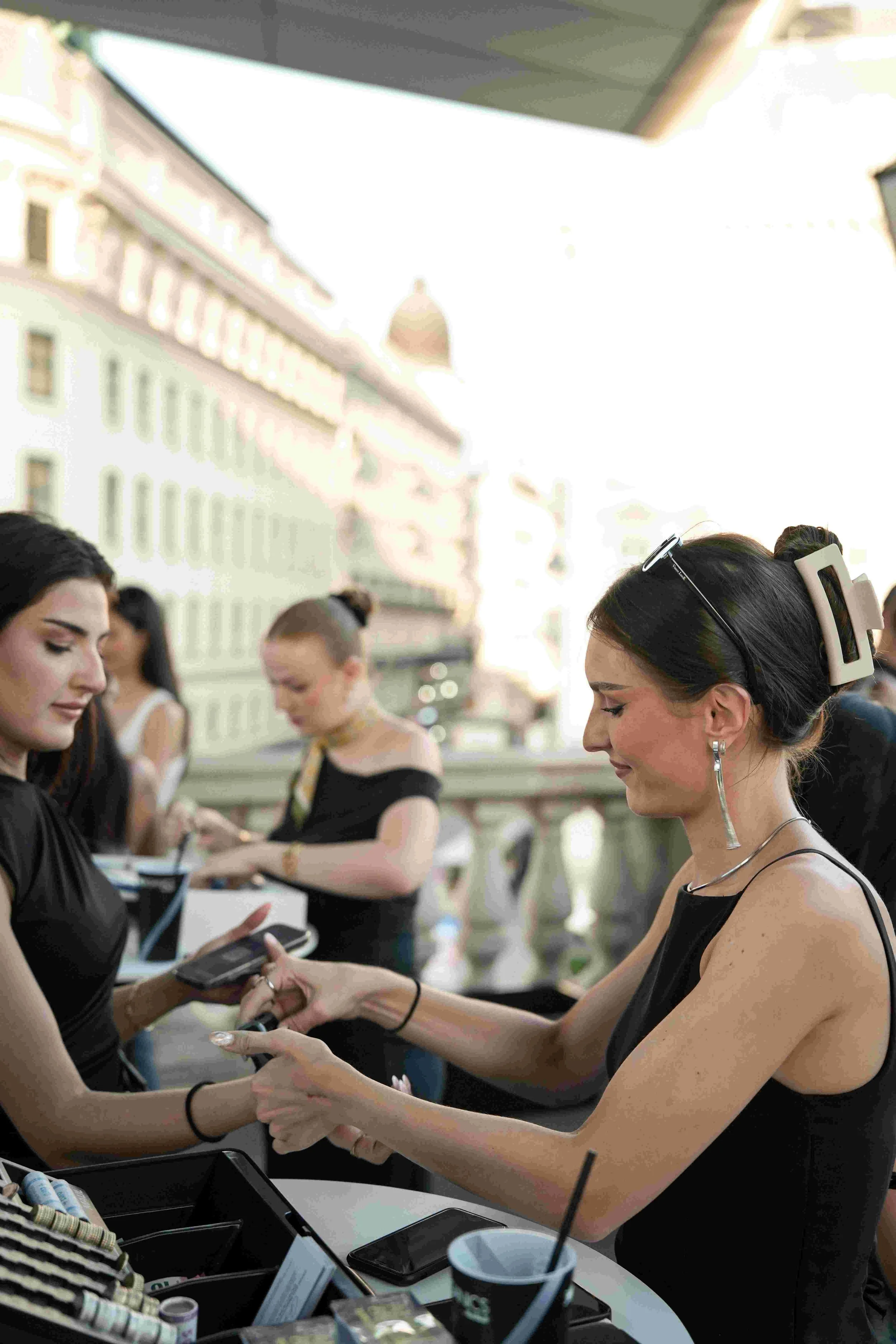 Women with dark hair and earrings at a registration or checkout table in an outdoor urban setting, interacting with a tablet or sign-in sheet, with several other women and buildings in the background.