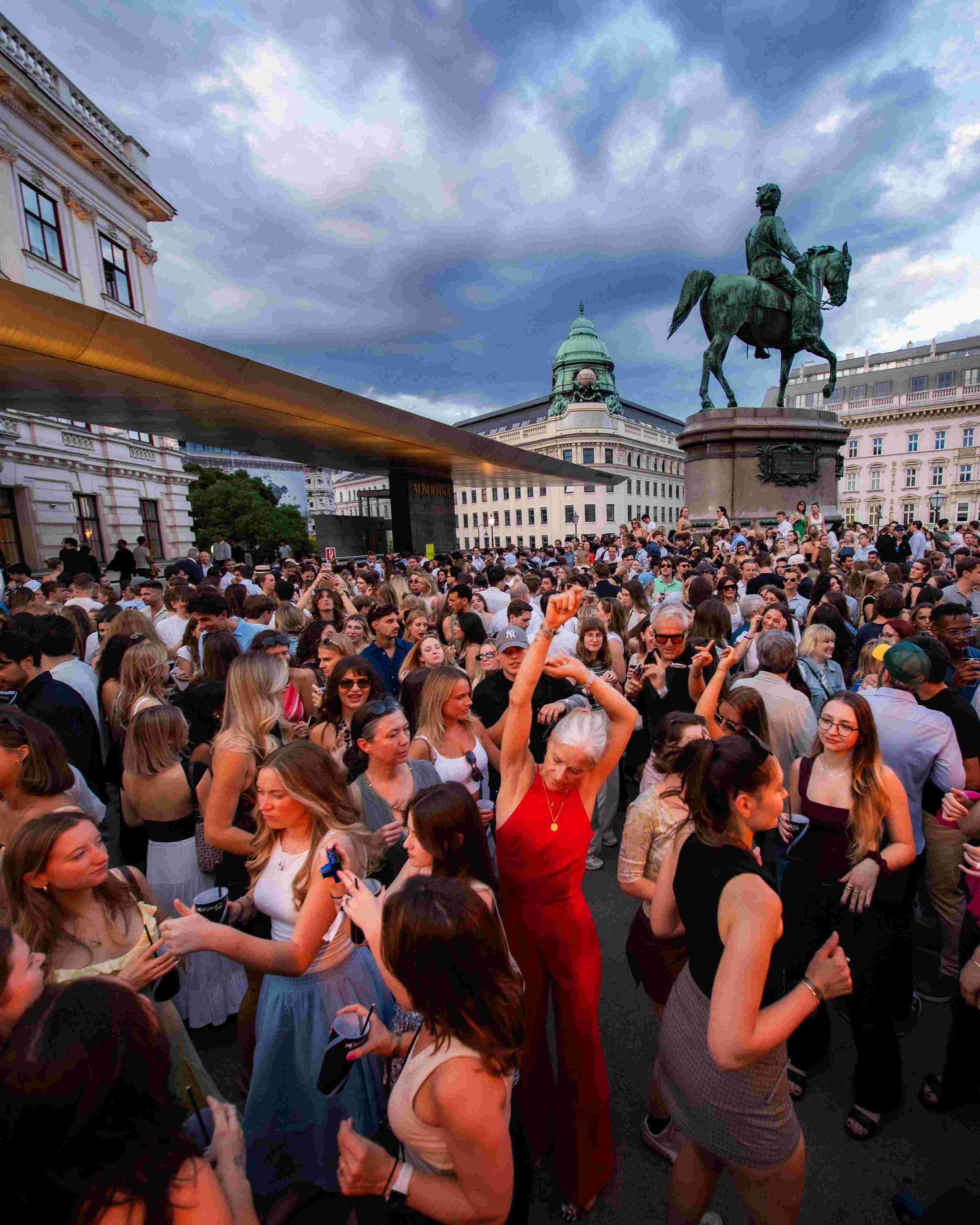 Crowd of people gathered outdoors in front of historic buildings and a statue of a man on horseback, under a cloudy sky.