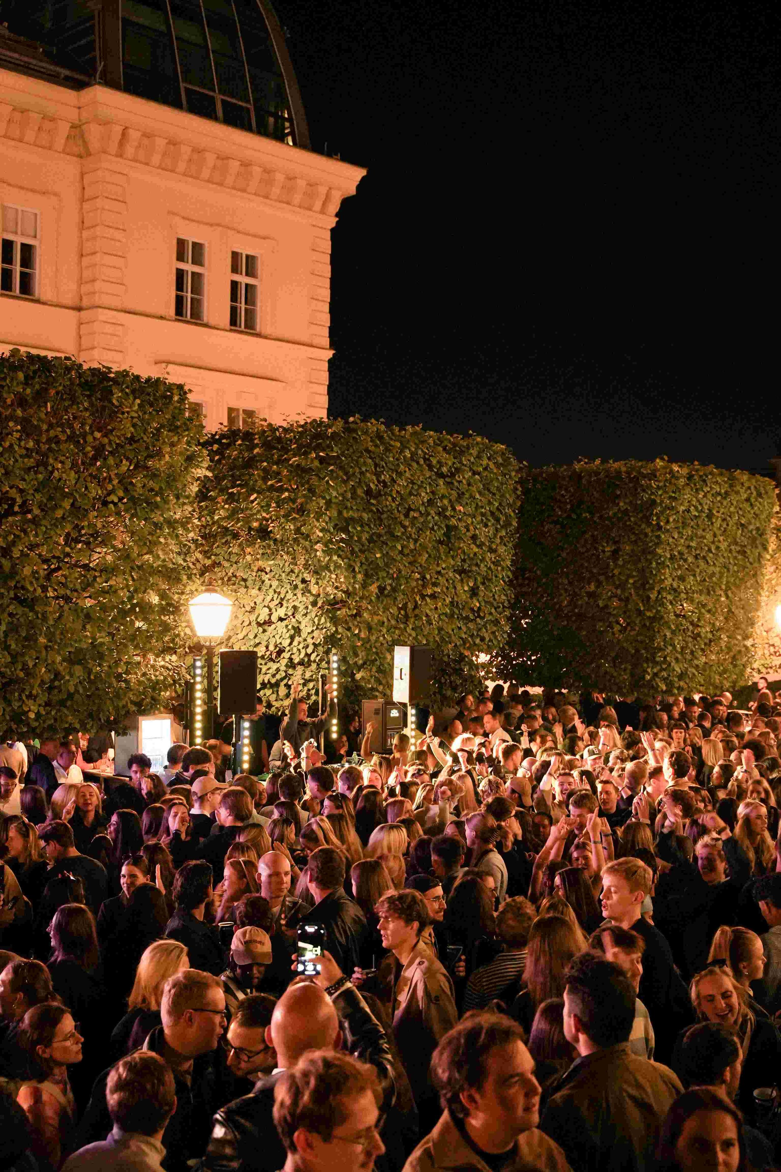 Crowd of people gathered outdoors at night, surrounded by tall trees and buildings, some taking photos, with street lamps illuminating the scene.