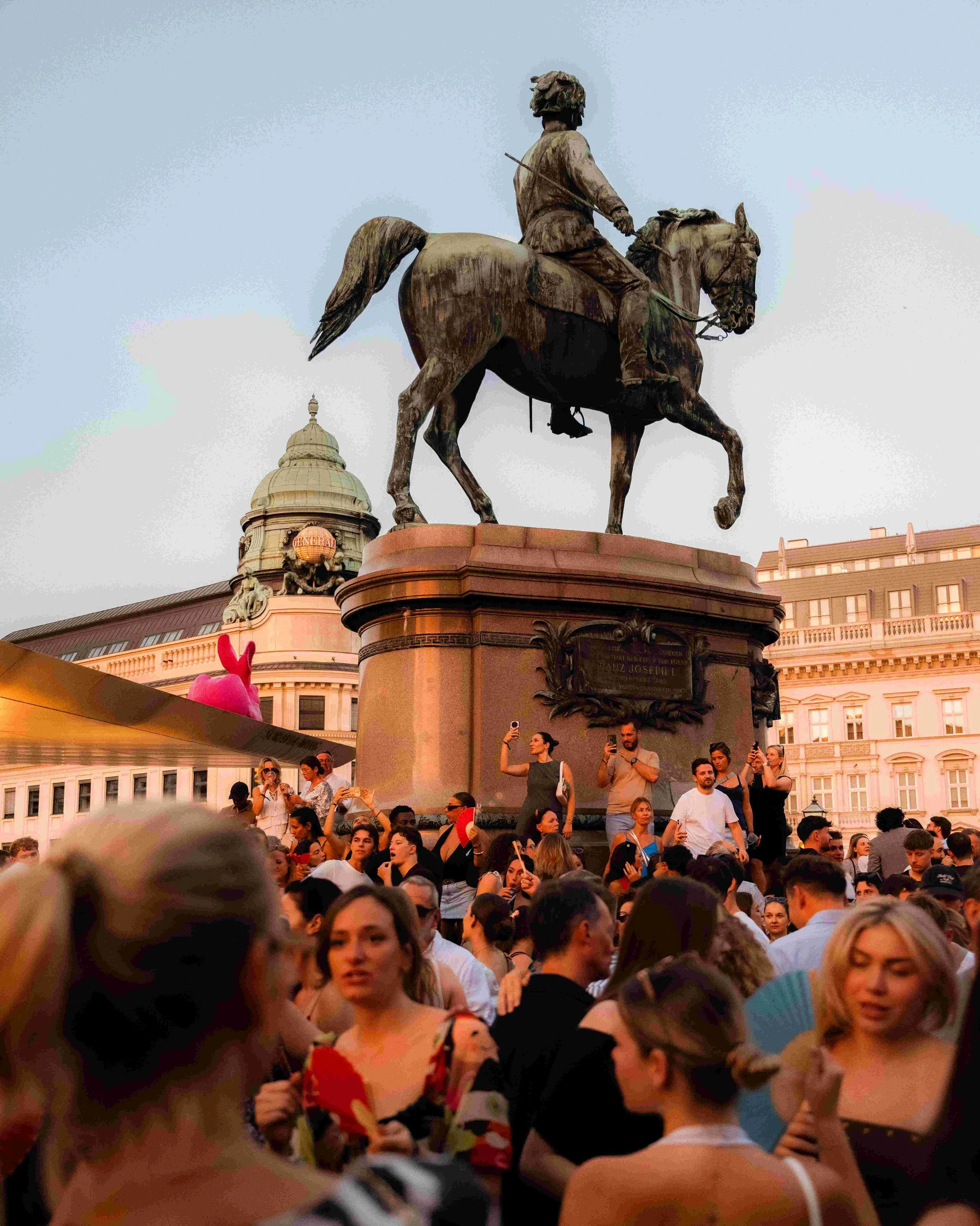 A large crowd of people gathered in front of a statue of a man on a horse in an urban square during daytime. The crowd appears diverse, with individuals taking photos and socializing. The background includes historic buildings with ornate architectur