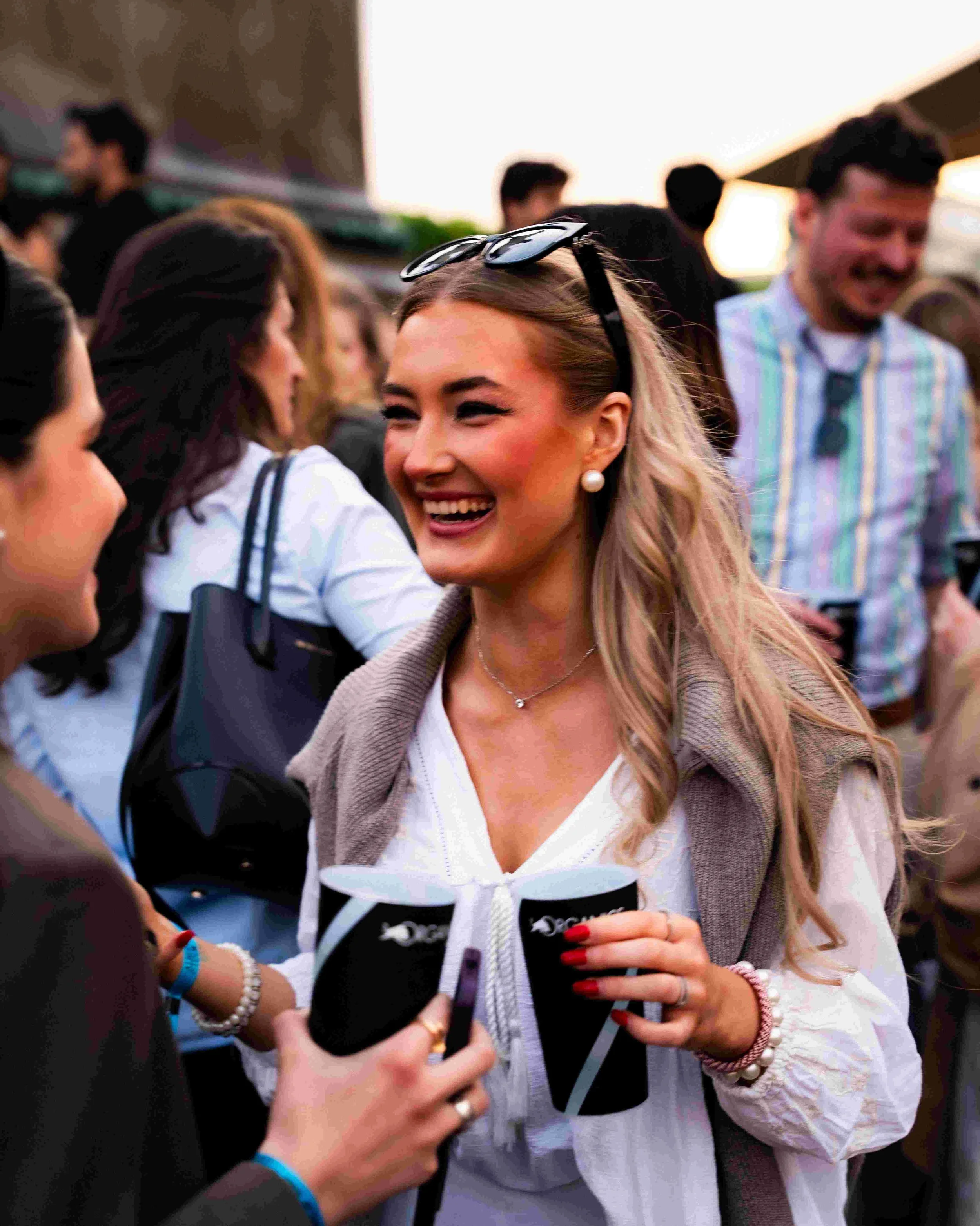 A smiling woman with blonde hair and a pearl earring, wearing sunglasses on her head, is looking at her phone while others around her are engaged in conversation.