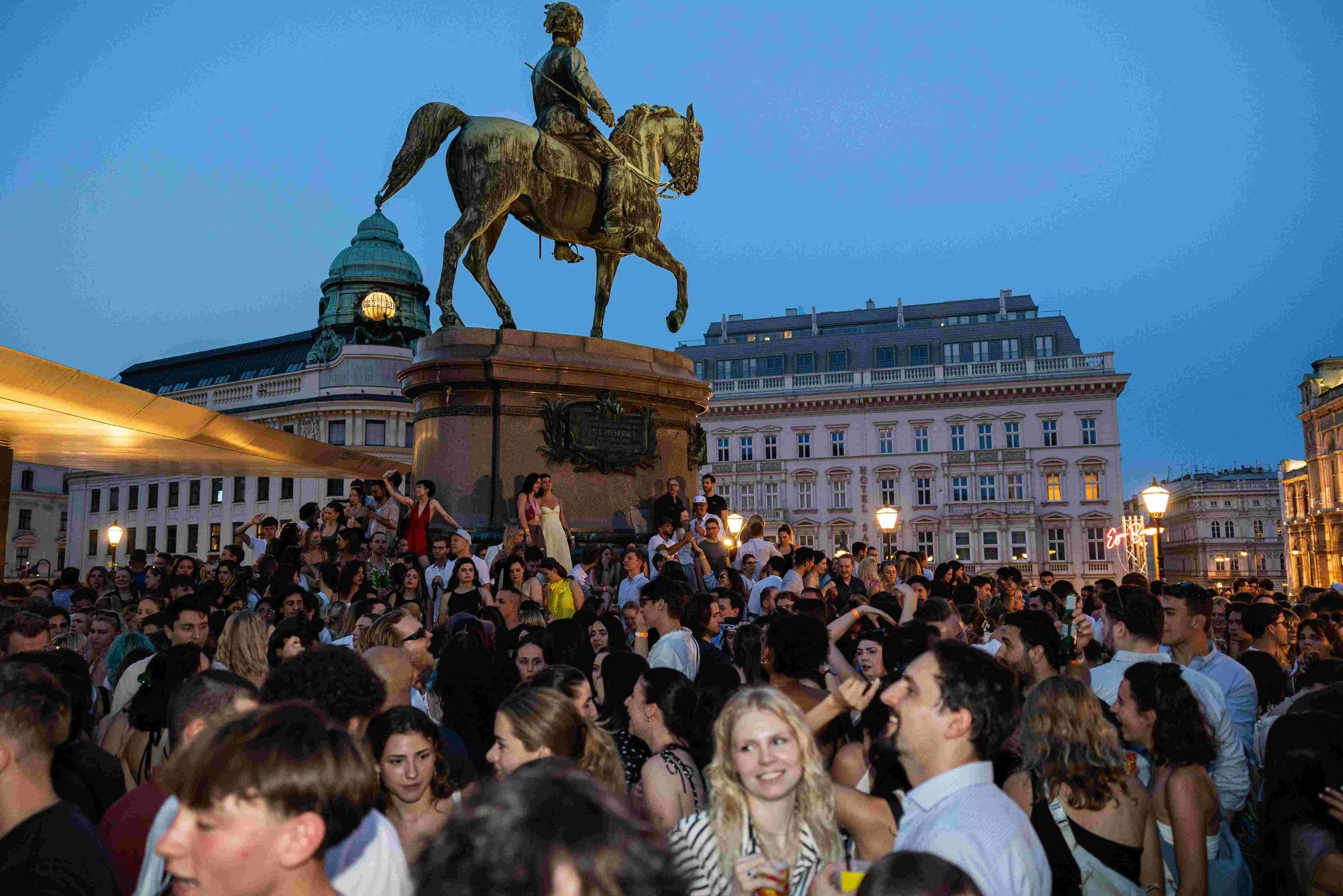 A crowded crowd of people gathered in front of a historic building with an equestrian statue on top, during dusk or evening.