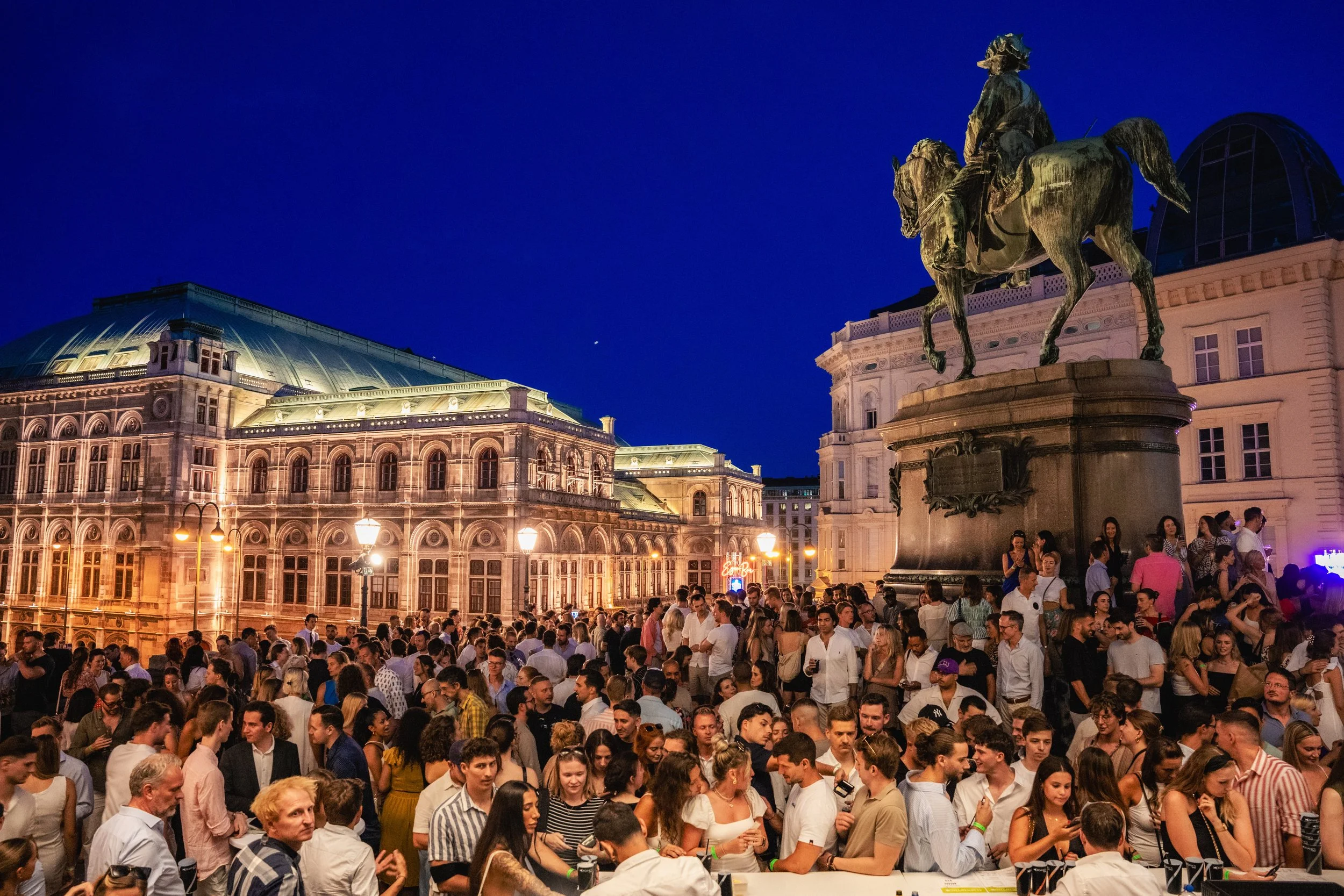 A large crowd of people gathered in a city square at night, with historic buildings and an equestrian statue under a dark blue sky.