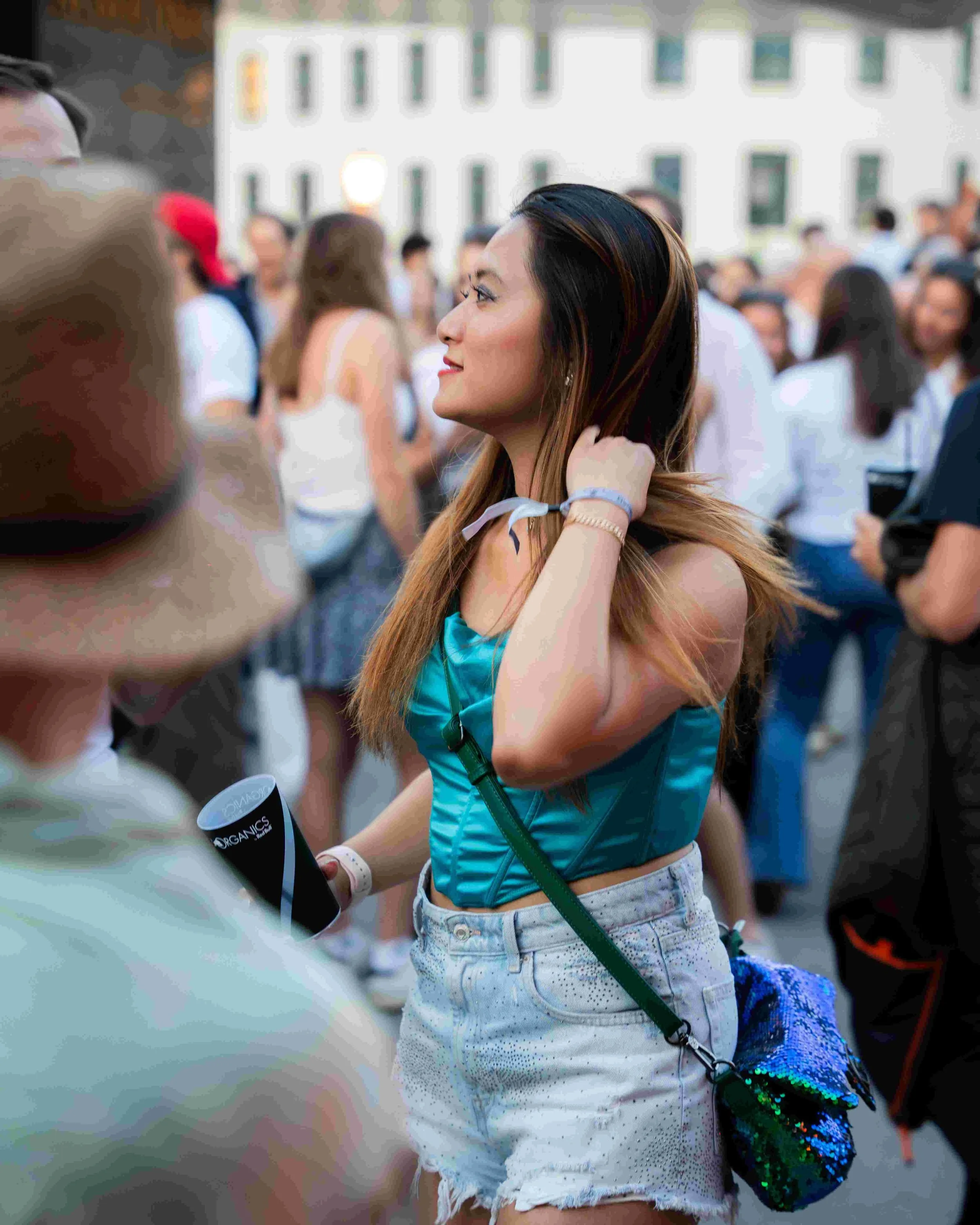 A young woman with long dark hair and a teal tank top adjusting her hair while holding a black tumbler in a crowd of people at an outdoor event.