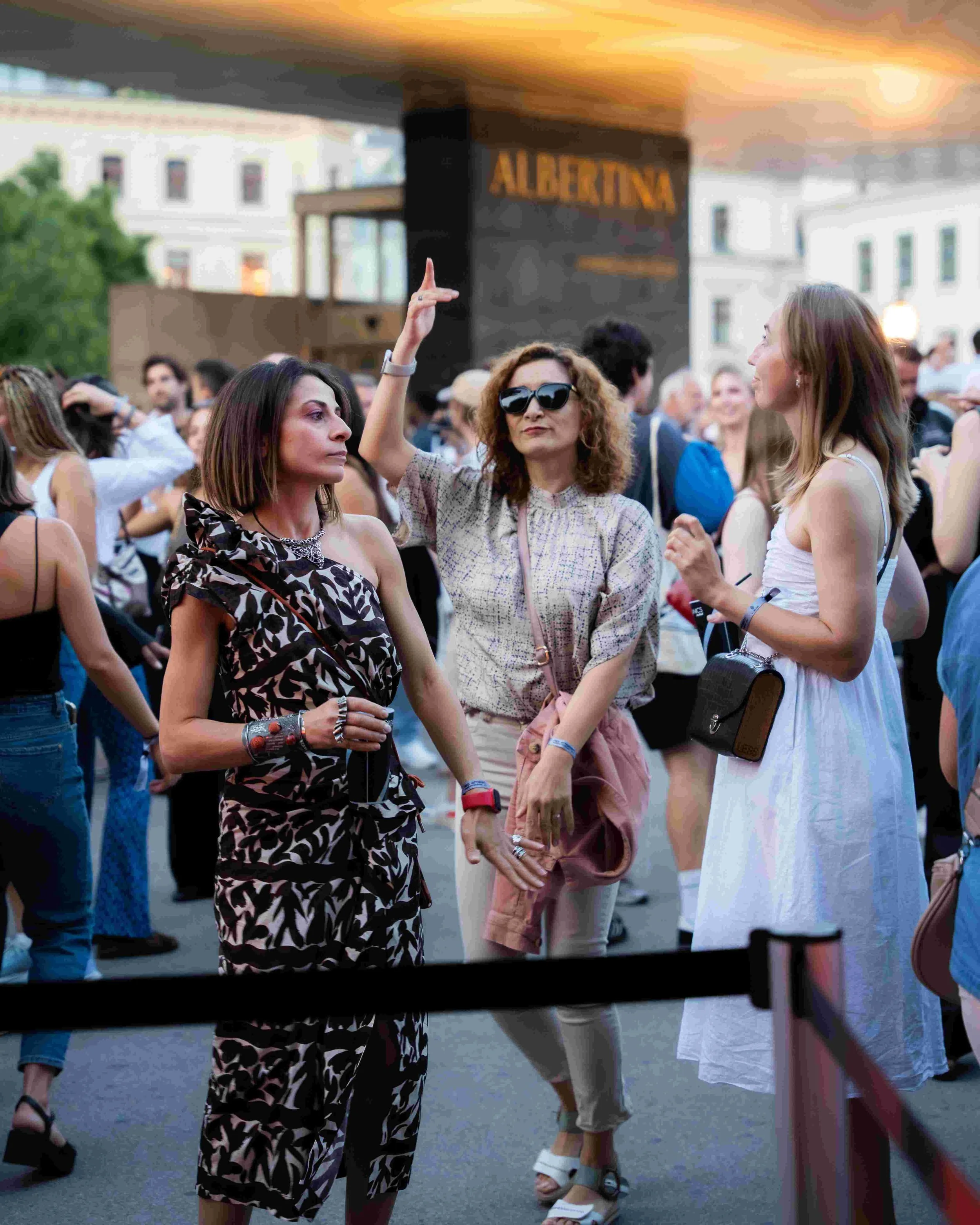 Group of women at outdoor event, including woman with curly hair and sunglasses, woman in white dress, and woman in patterned dress, with a barricade and sign in the background.