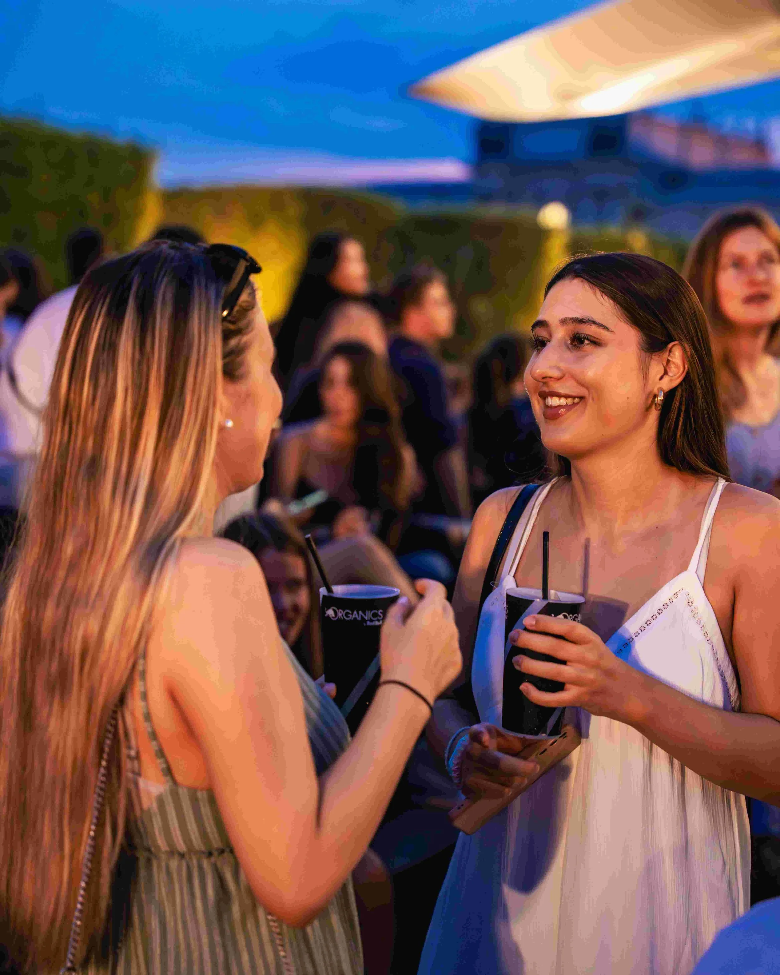 Two women at an outdoor evening event holding drinks and talking, with other people in the background.