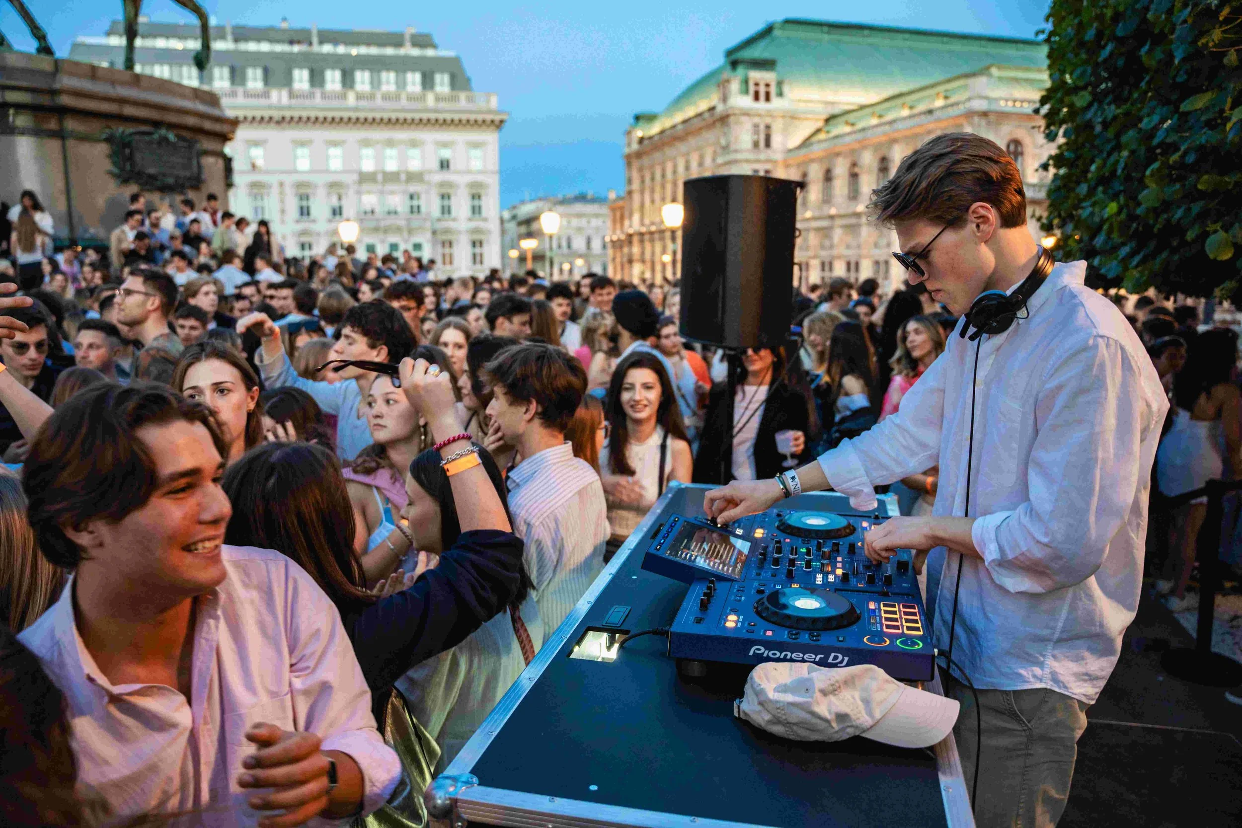 A DJ in sunglasses and white shirt playing music at an outdoor event during dusk, surrounded by a large crowd of young people in front of historic buildings.