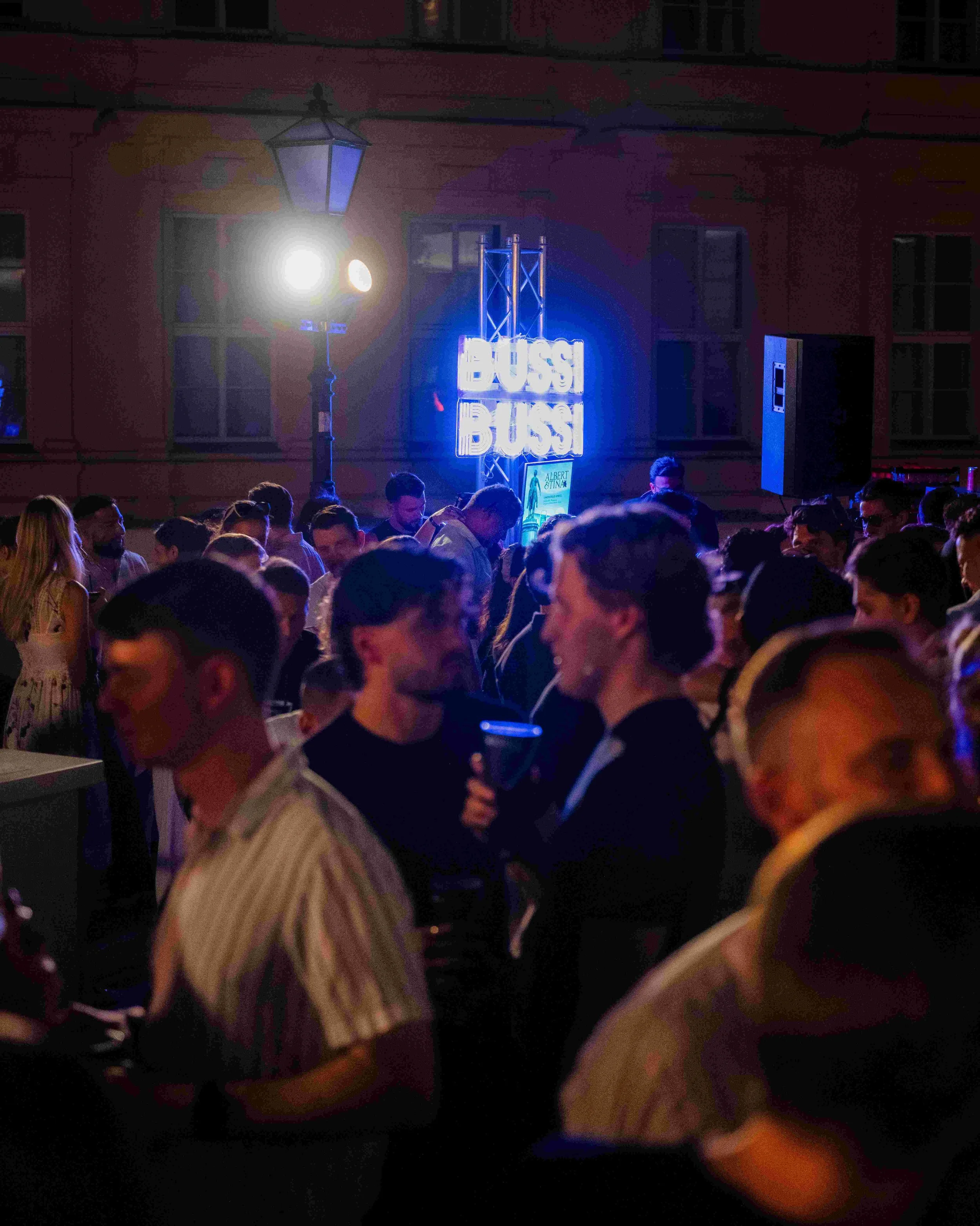 Nighttime scene at an outdoor event with a lively crowd, a bright neon sign reading 'BOSS BASS,' a lit street lamp, and a pink building facade with windows.