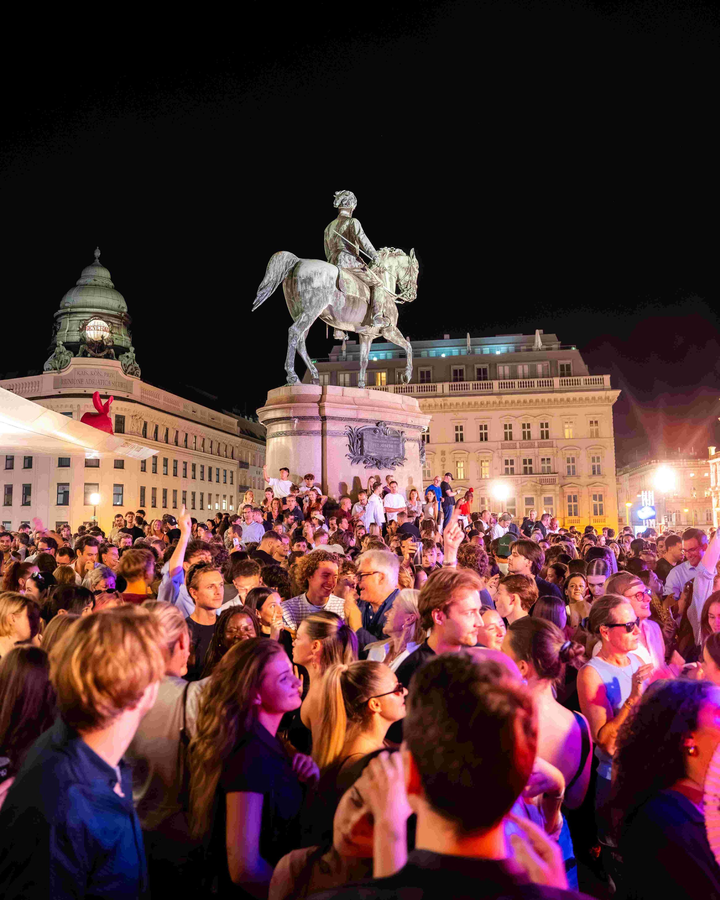 Nighttime scene of a large crowd gathered in a square with a statue of a man on horseback at the center. The crowd is diverse, with some people smiling and talking. The surrounding buildings are lit up, and the sky is dark.