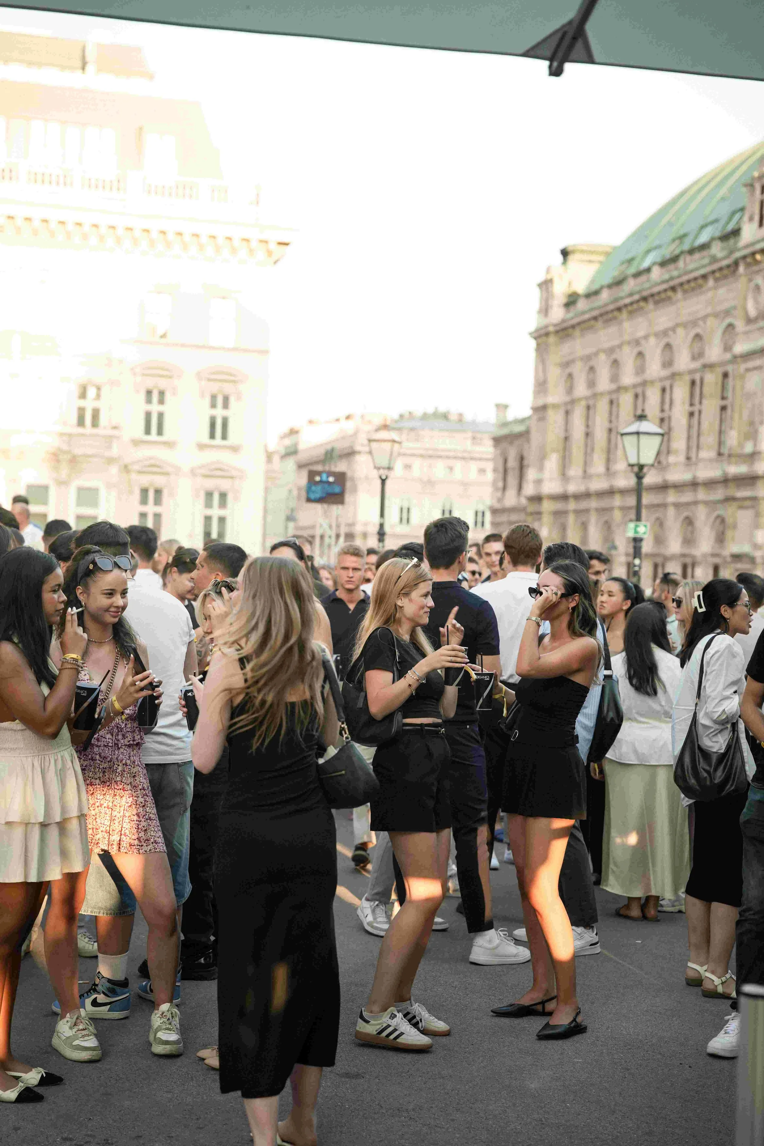 A crowd of people gathered outdoors in front of historic European-style buildings, some taking photos and engaging in conversation.