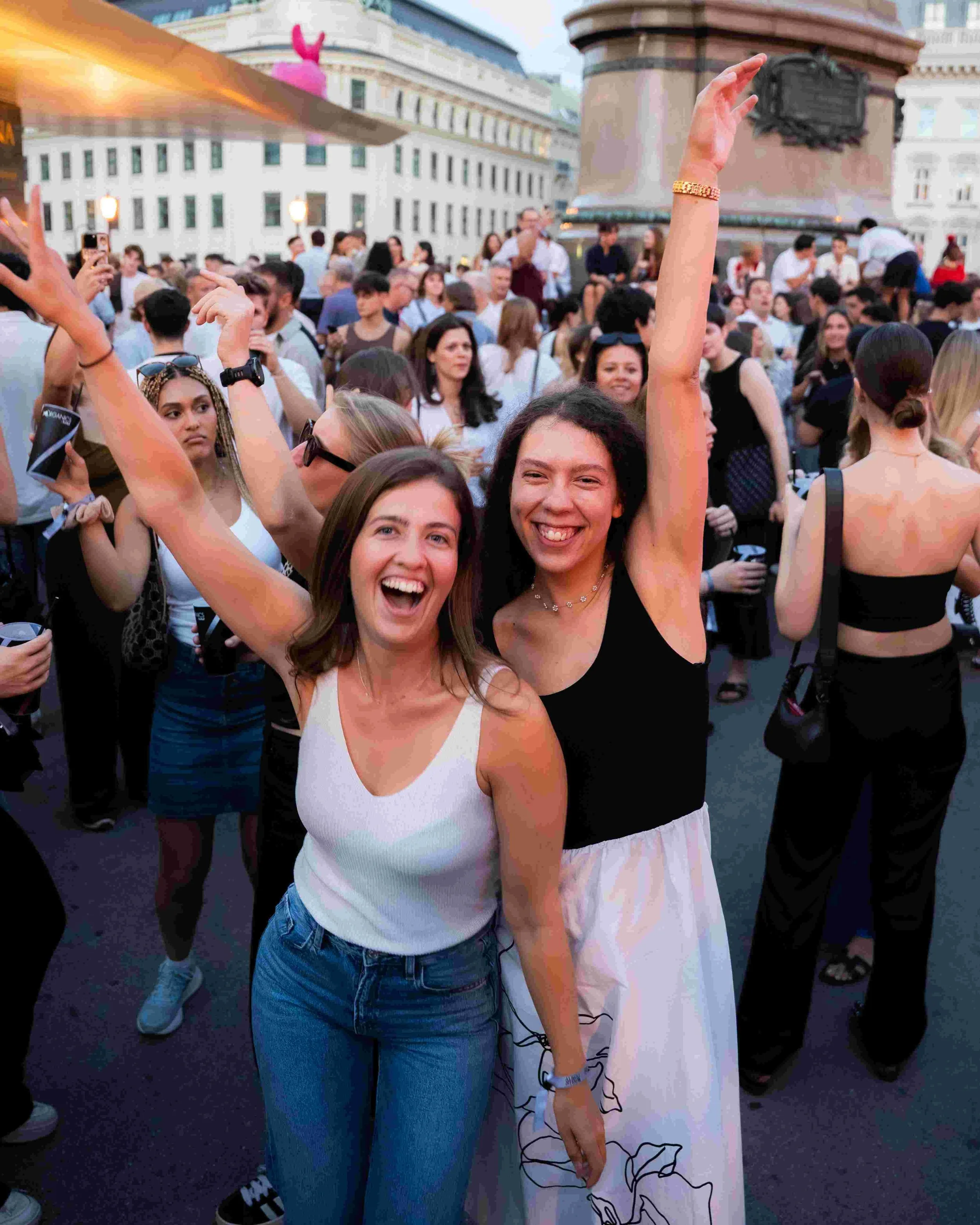 Two young women smiling and posing in a crowd at an outdoor event, with buildings and a large statue in the background.