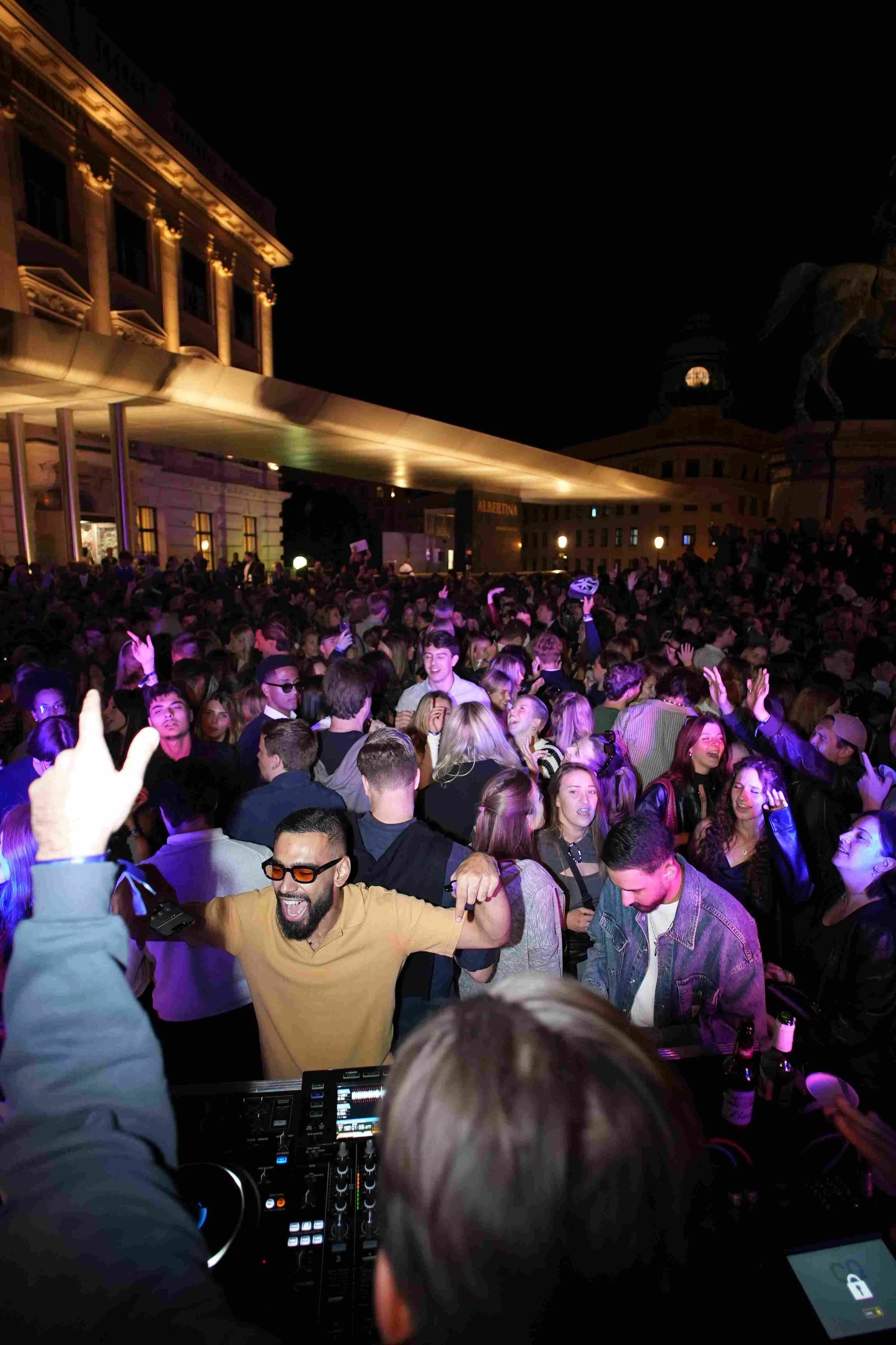 Crowd of people at a nighttime outdoor event, dancing and enjoying music, with some raising their hands and smiling.