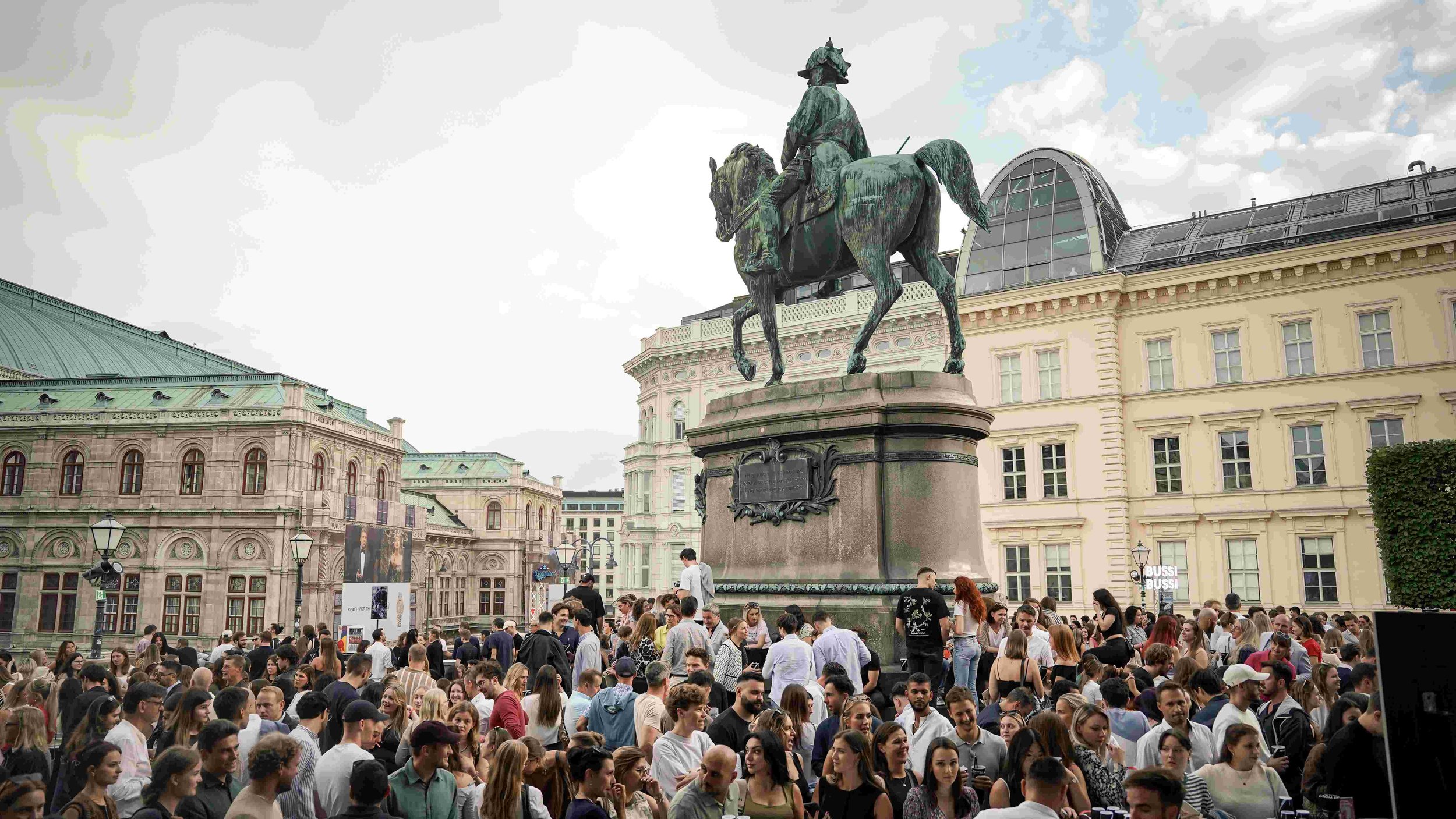 Crowd of people gathered around an equestrian statue of a historical figure in a city square, with classical and modern buildings in the background.