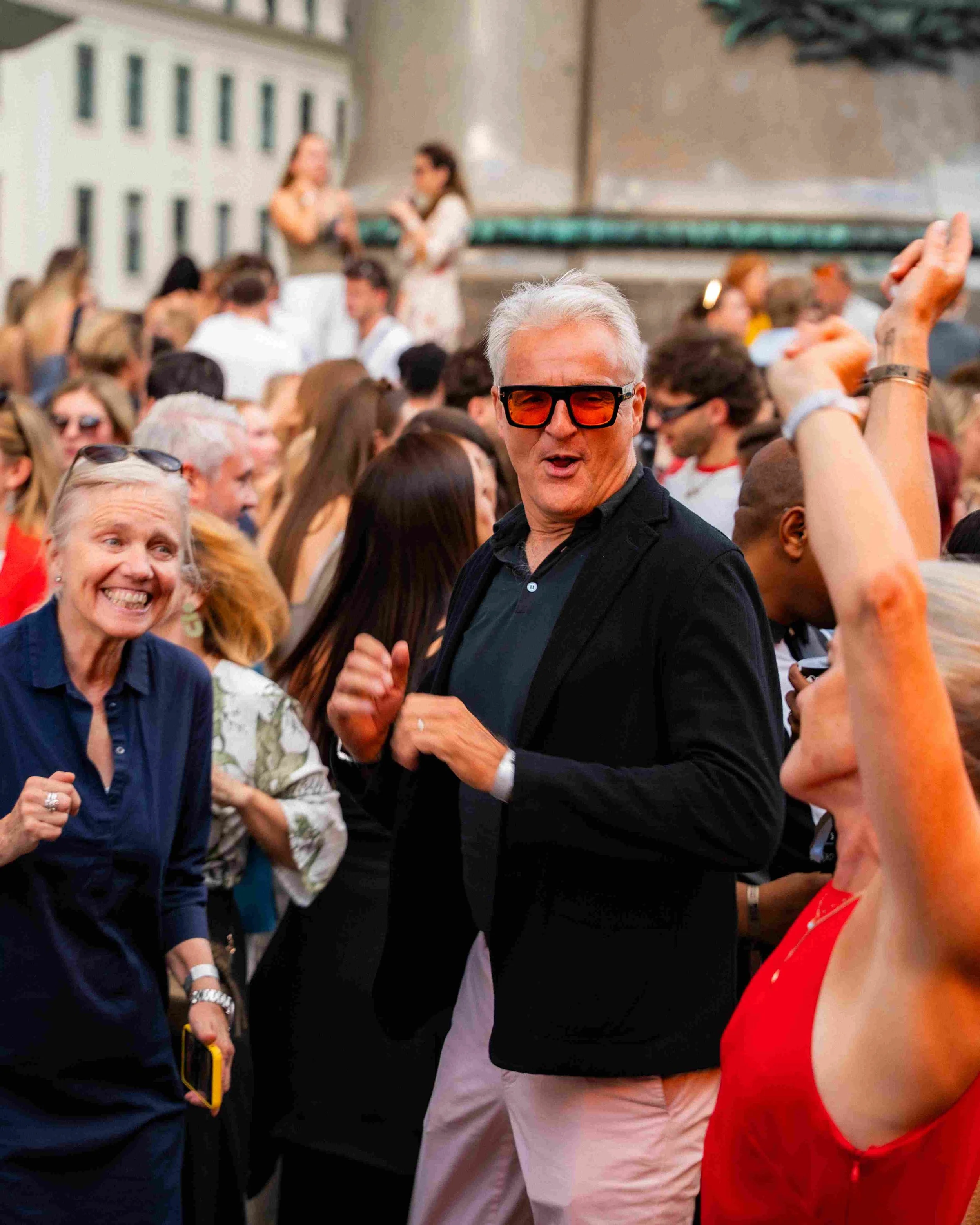 A crowd gathered outdoors with two women talking in the background. The focus is on a man with gray hair, wearing large sunglasses, a black blazer, and a dark shirt, who is smiling and interacting with others. A woman in a red dress is partially visi