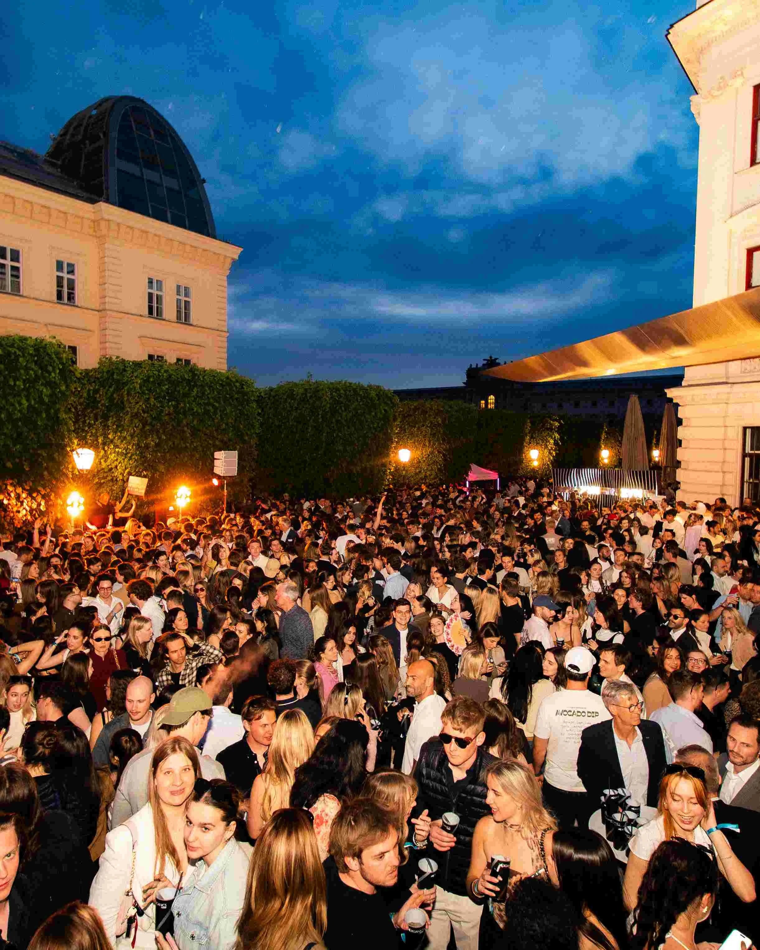 A large crowd of people gathered outdoors during the evening, with some holding drinks and engaging in conversations. The background features buildings and darkening sky.