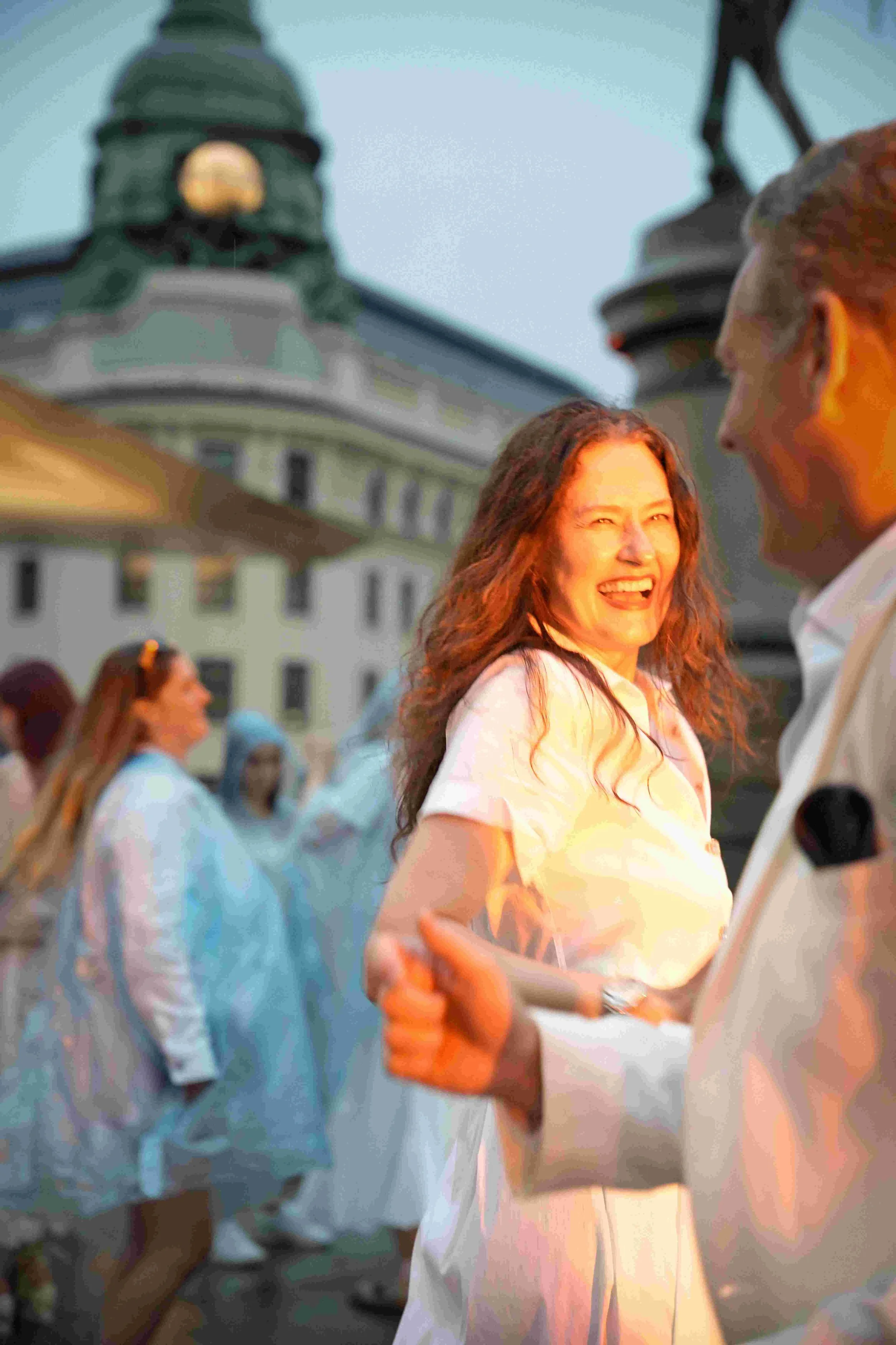 A group of people outside, some are dancing and smiling. The photo focuses on a woman with long red hair who is smiling and wearing a light-colored dress, with other people around her and a historic building in the background.