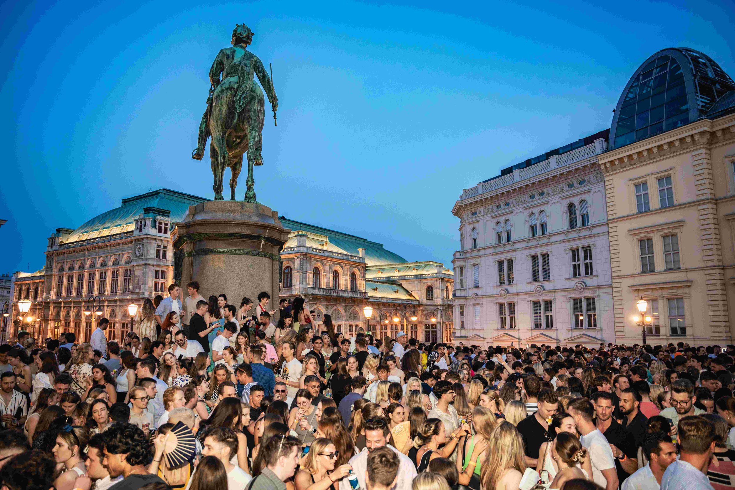 Crowd of people gathered outdoors in front of historic buildings and a statue of a man on a horse during dusk, with the lit city skyline in the background.