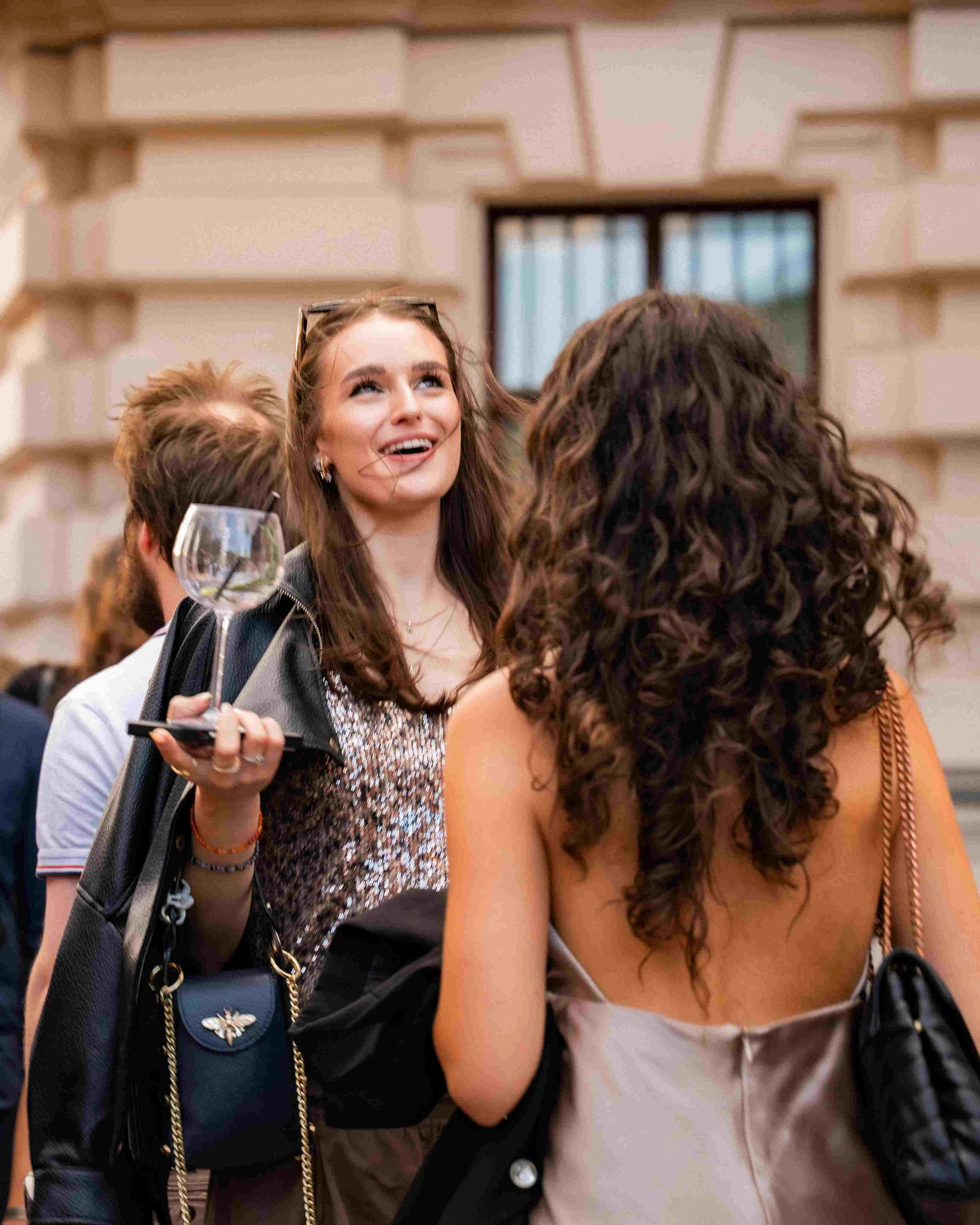 A woman with long brown hair and earrings smiling and looking up, holding a glass of wine, with other people around her in a social gathering indoors.