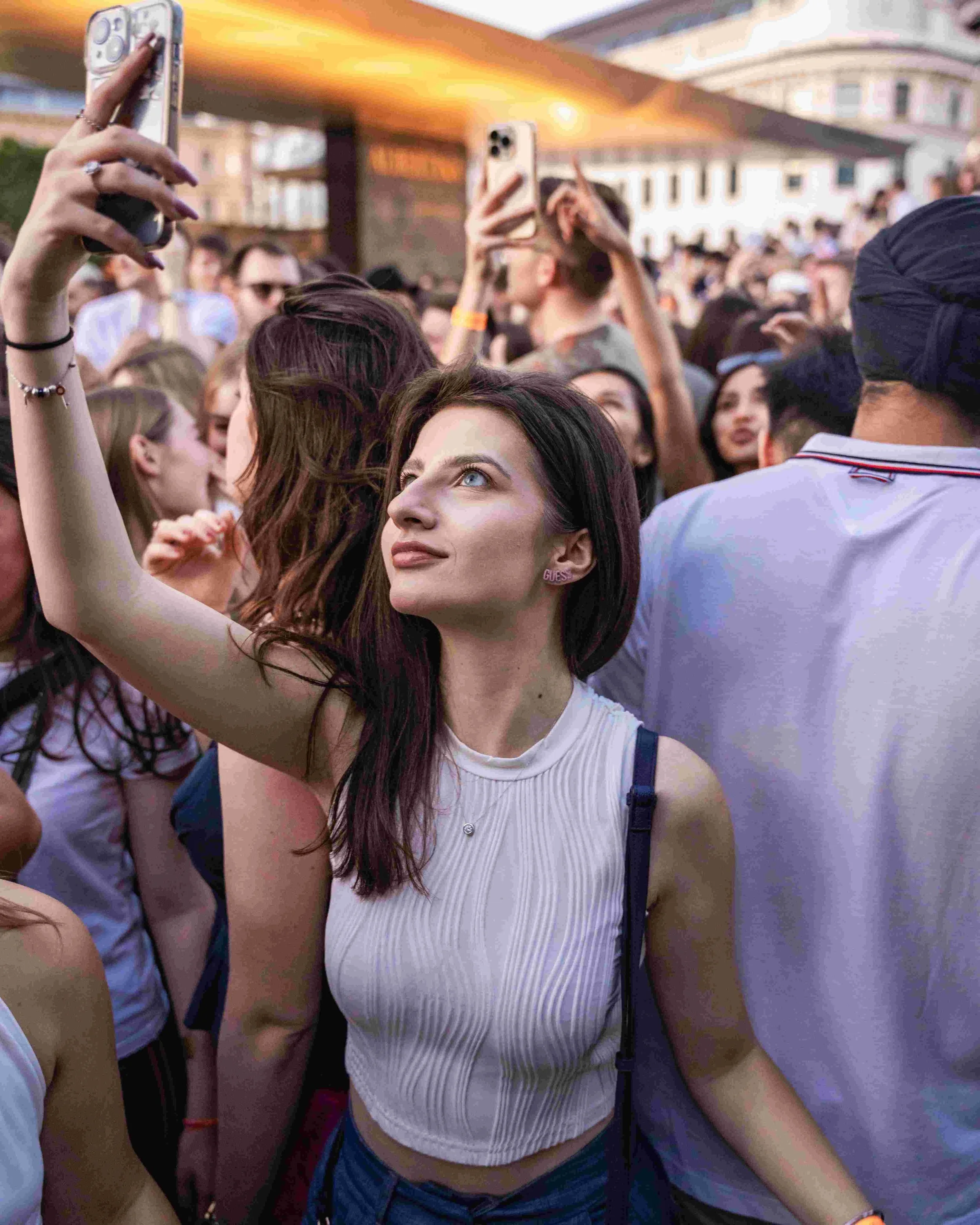 Young woman with dark hair taking a selfie at an outdoor event with a diverse crowd of people in the background. She is wearing a white sleeveless top and has a backpack.