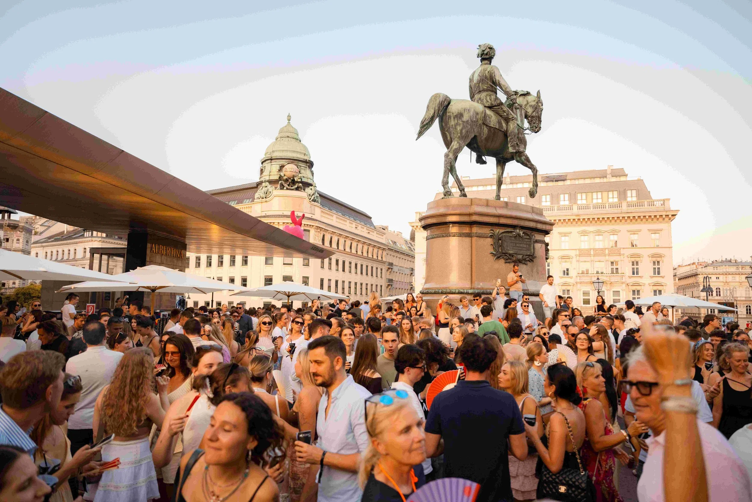 Crowd gathered in a city square with a large equestrian statue in the background and historic buildings around.