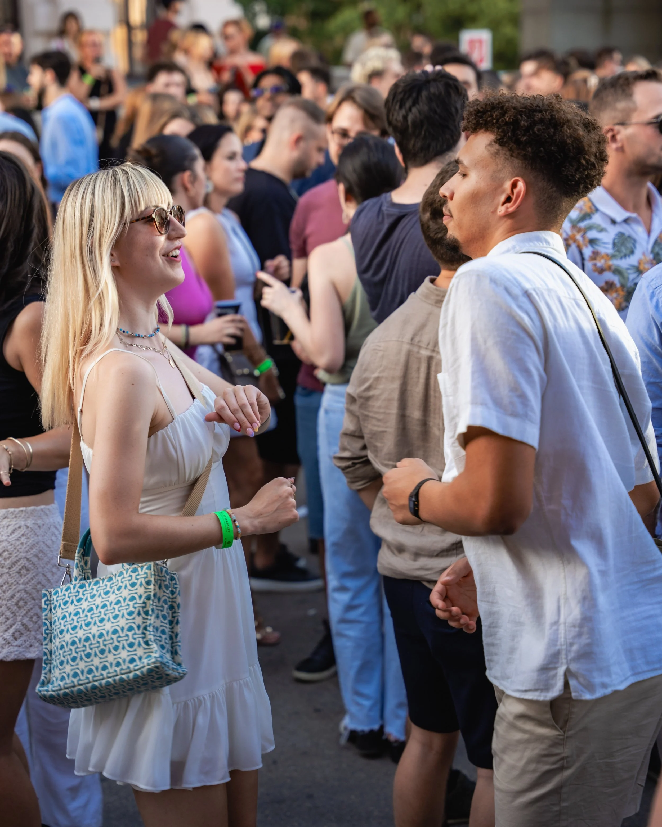 A group of people at an outdoor event, engaging in conversations, with a focus on a blonde woman wearing sunglasses and a white dress, and a man with curly hair and a white shirt. Many people are in the background.