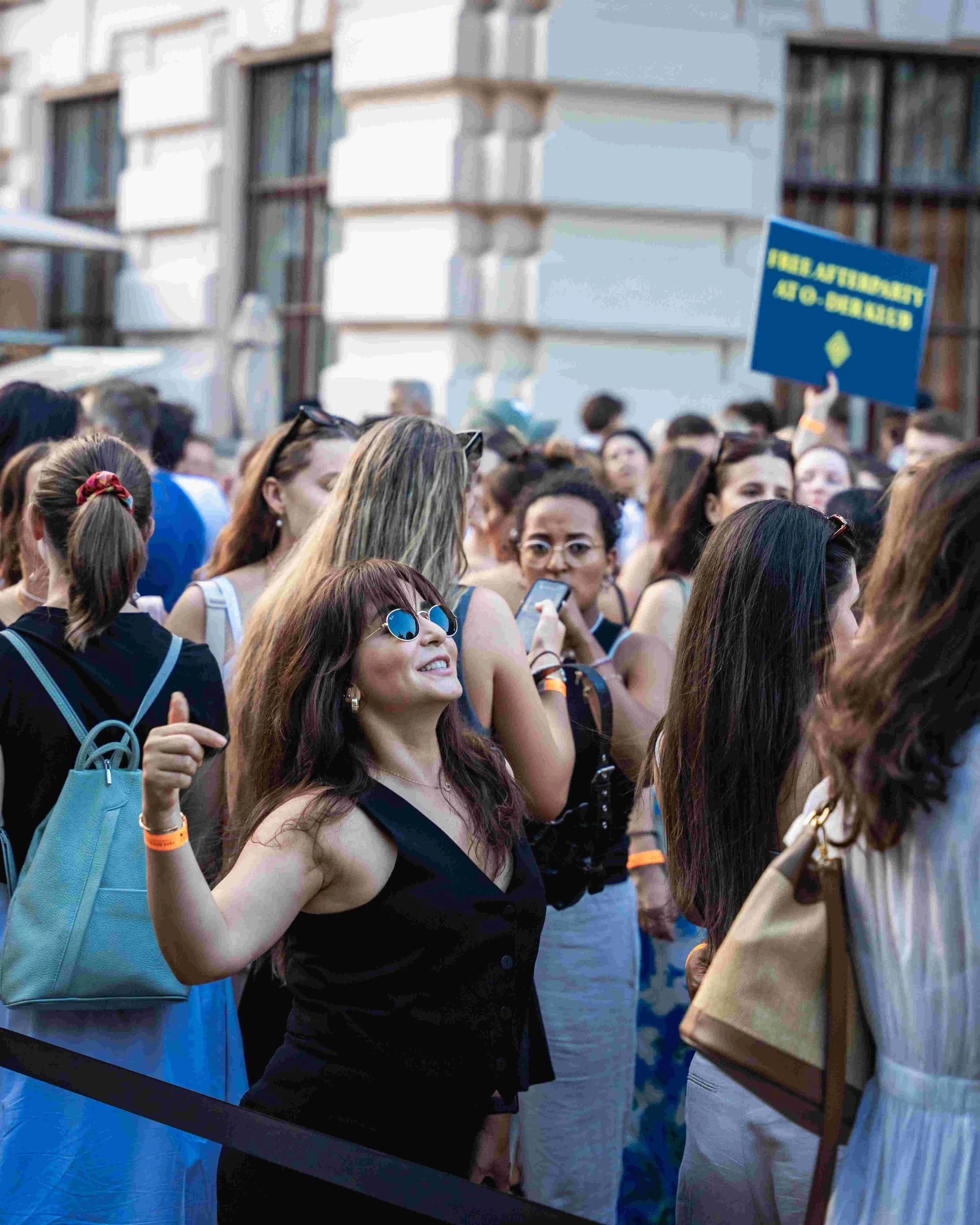 Crowd of people at an outdoor event or exhibit, some holding signs and taking photos, with a building in the background.