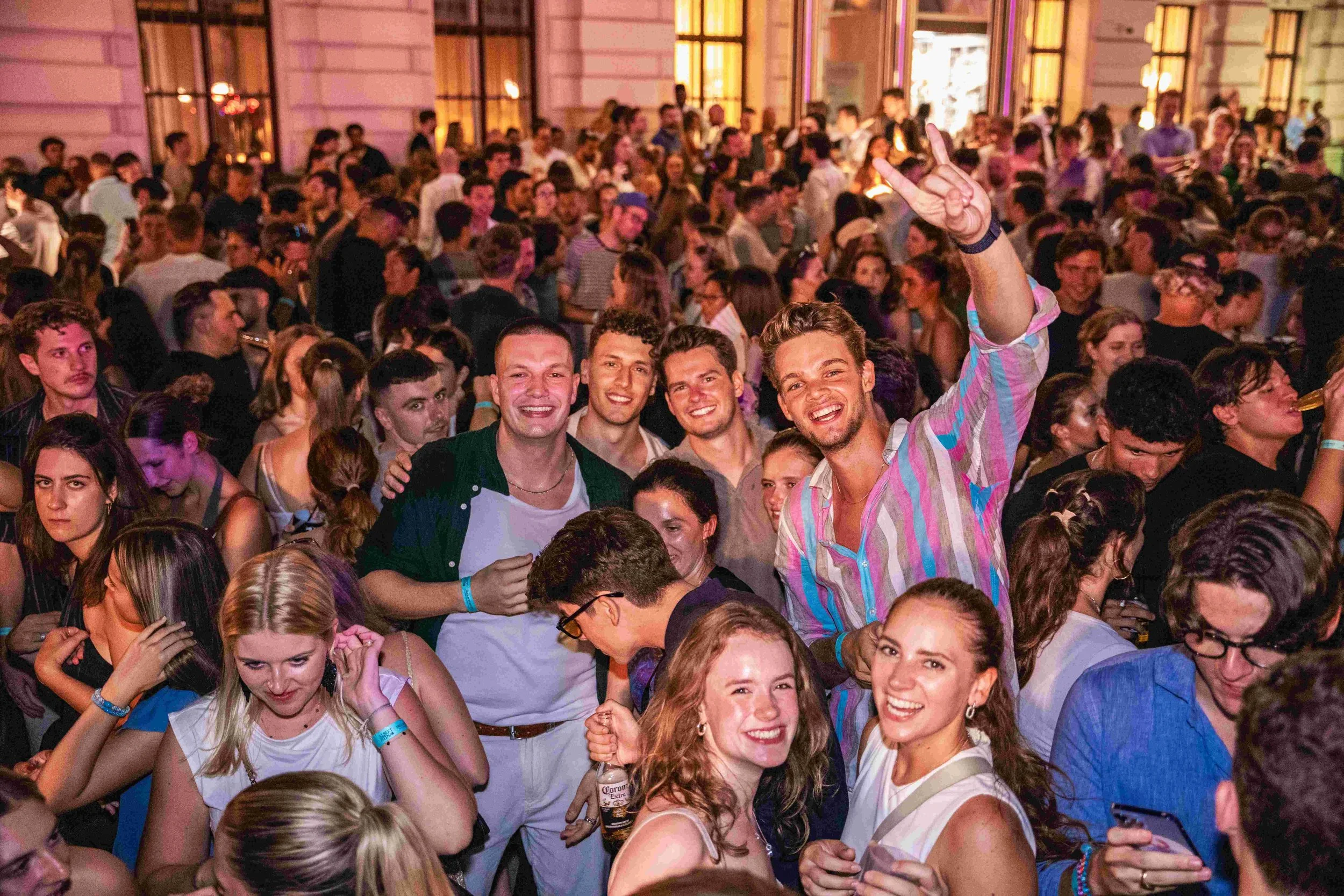 A large crowd of young people at a party or concert with many smiling and talking. The scene is indoors with warm, colorful lighting.