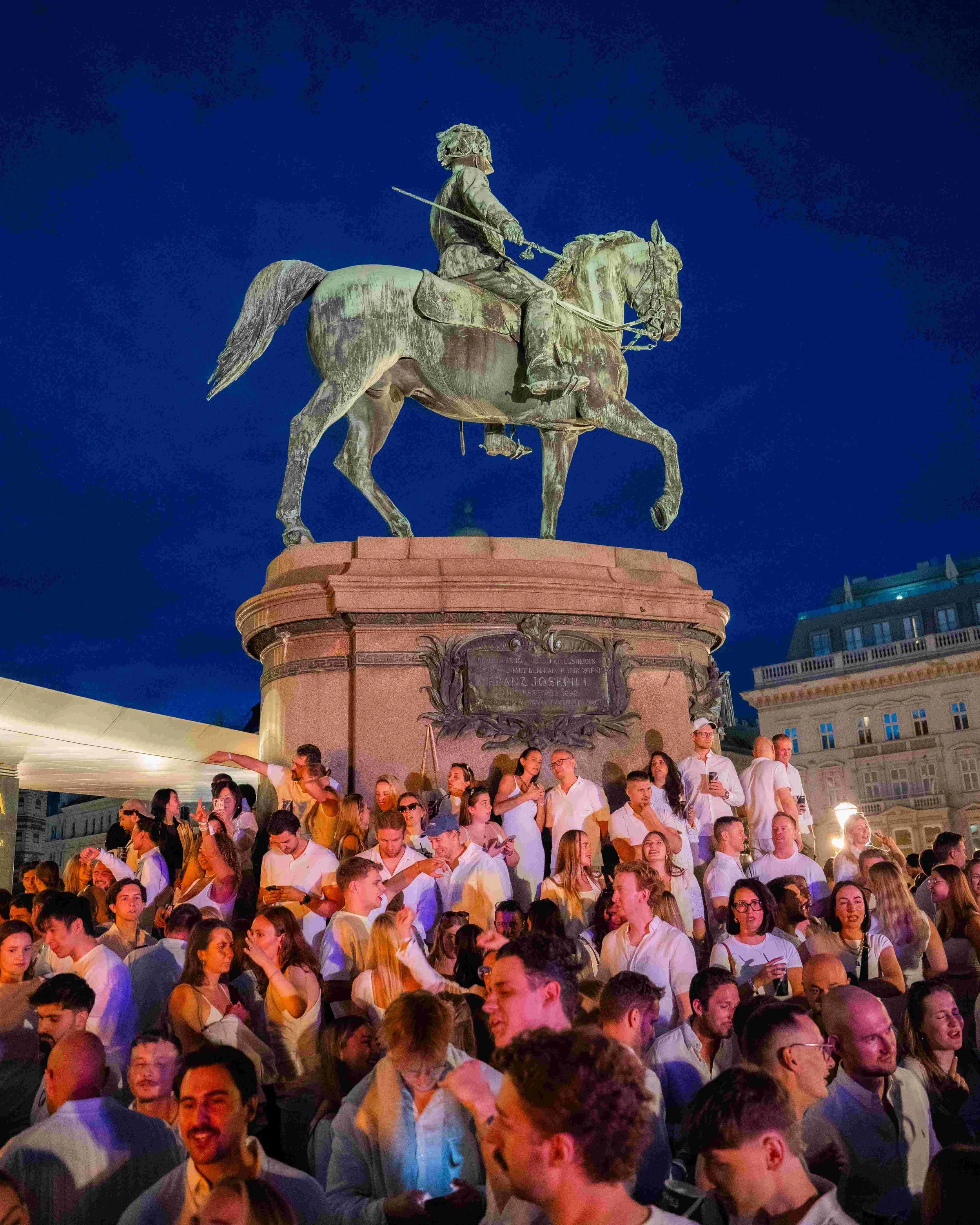 A large crowd of people gathered at night around a bronze equestrian statue of a man in military uniform holding a baton, mounted on a pink stone pedestal. The scene is illuminated with artificial lighting against a dark blue sky.